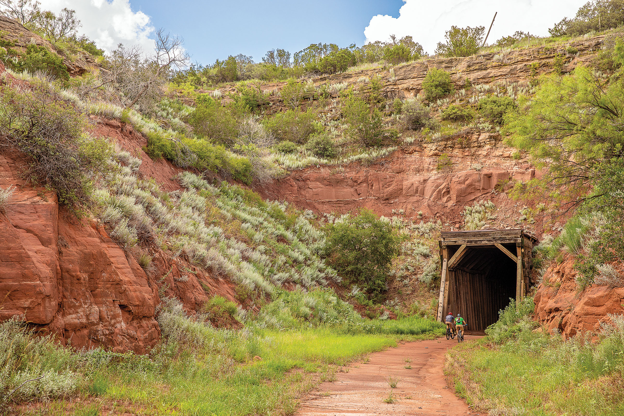 Two people ride bicycles toward the entracnce of a large tunnel inside a red and green hillside