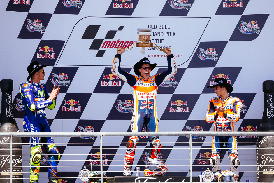Marc Marquez celebrating his victory on the podium, raising his vaguely longhorn-shaped trophy above his head.