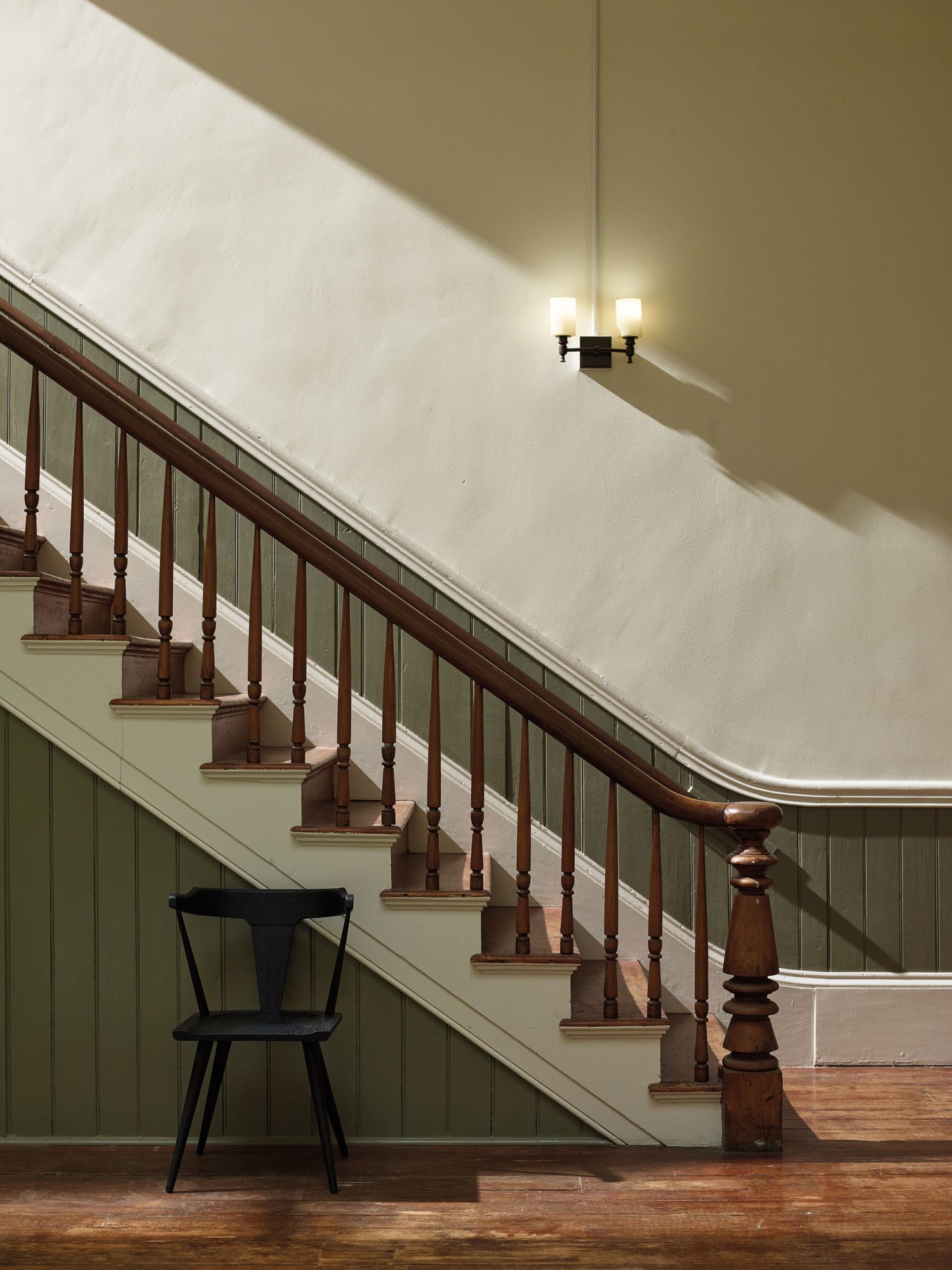 A single light fixture casts long shadows along a plain white wall behind an ornate wooden staircase