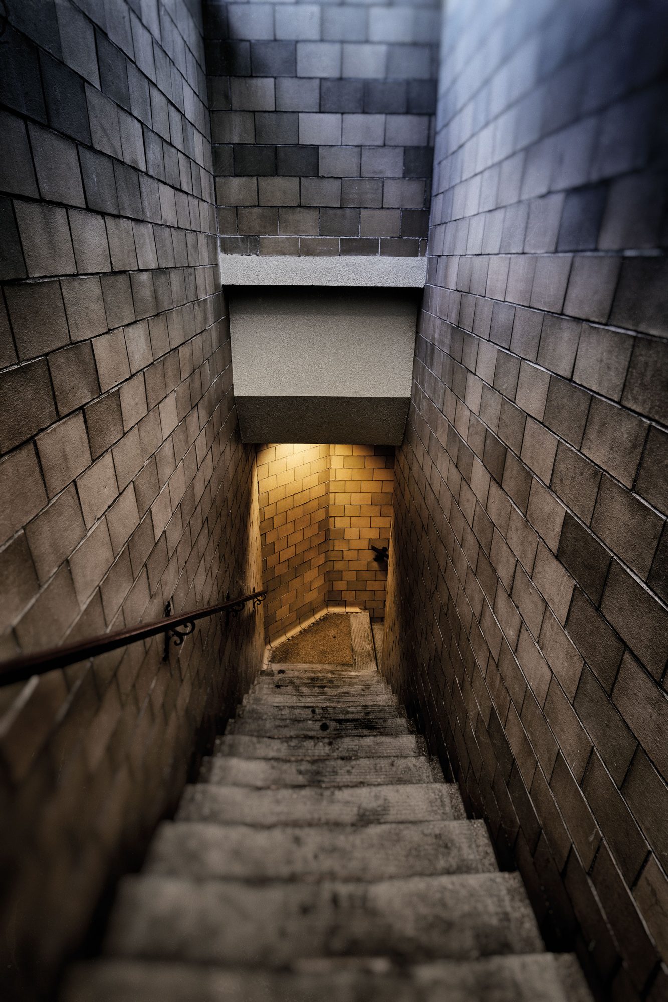 Tall concrete-brick walls line a gray staircase down to a room illuminated by a yellow light