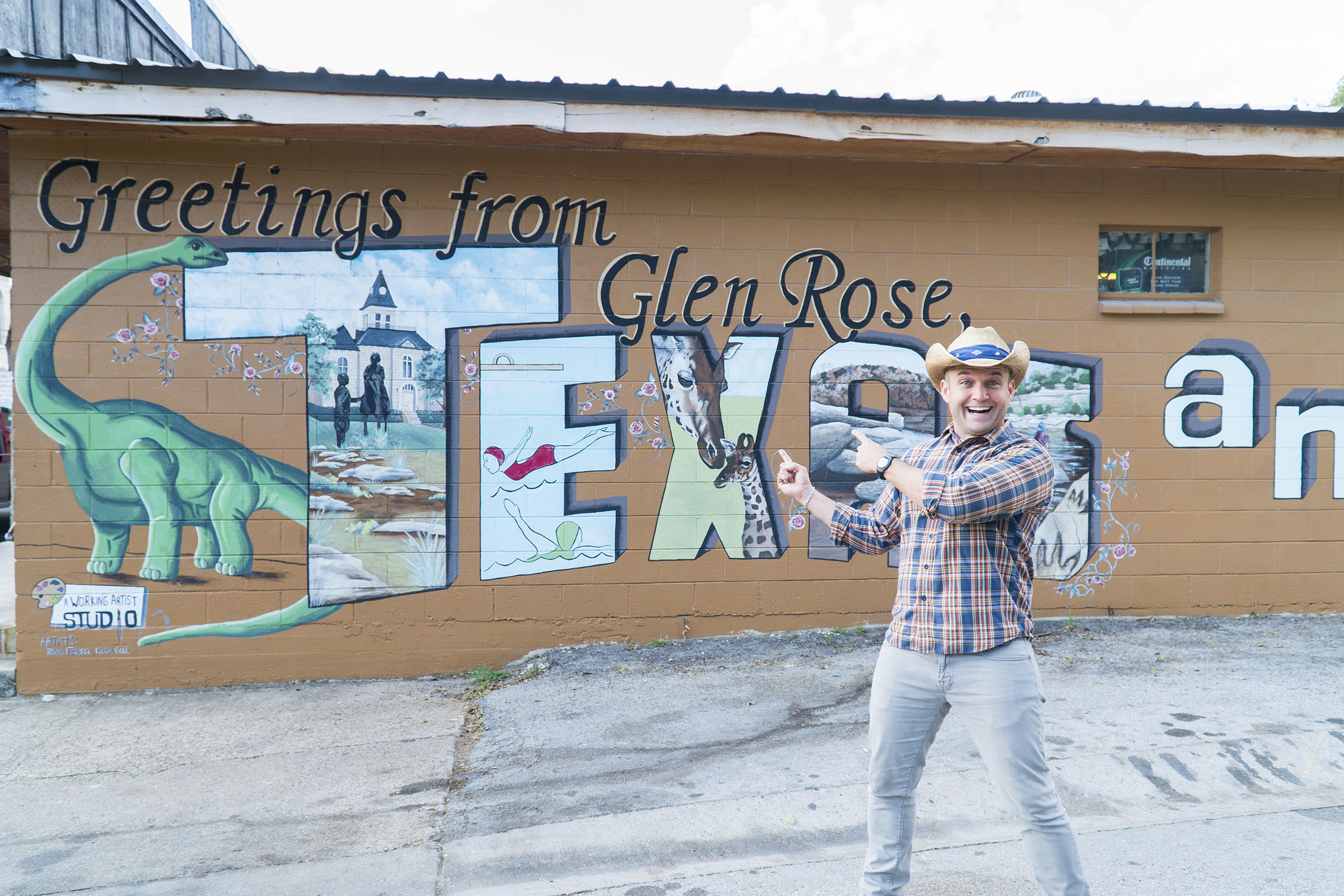 The Daytripper, Chet Garner, poses outside of a mural featuring a dinosaur that says "Greetings from Glen Rose Texas"