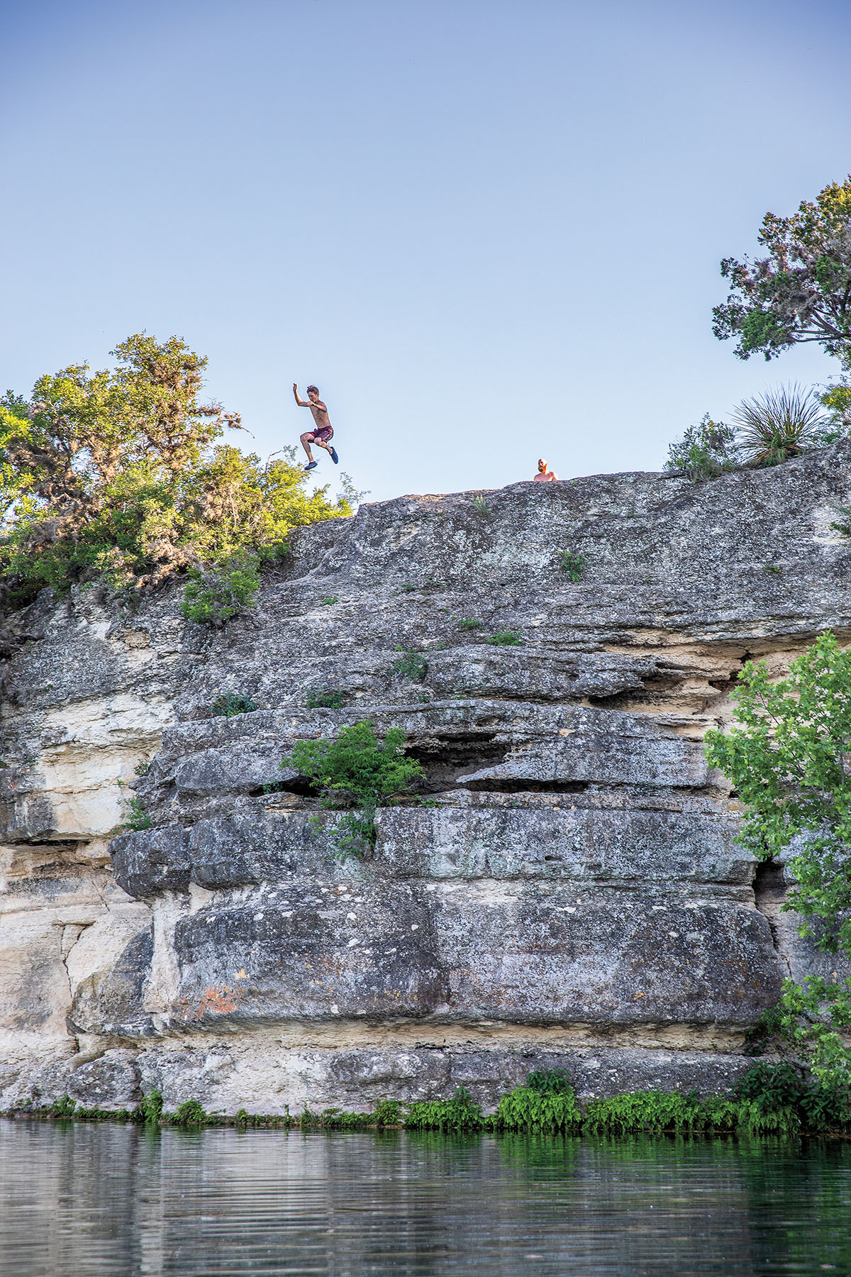 Young men jump off of a rock cliff into the water of Lake Nueces