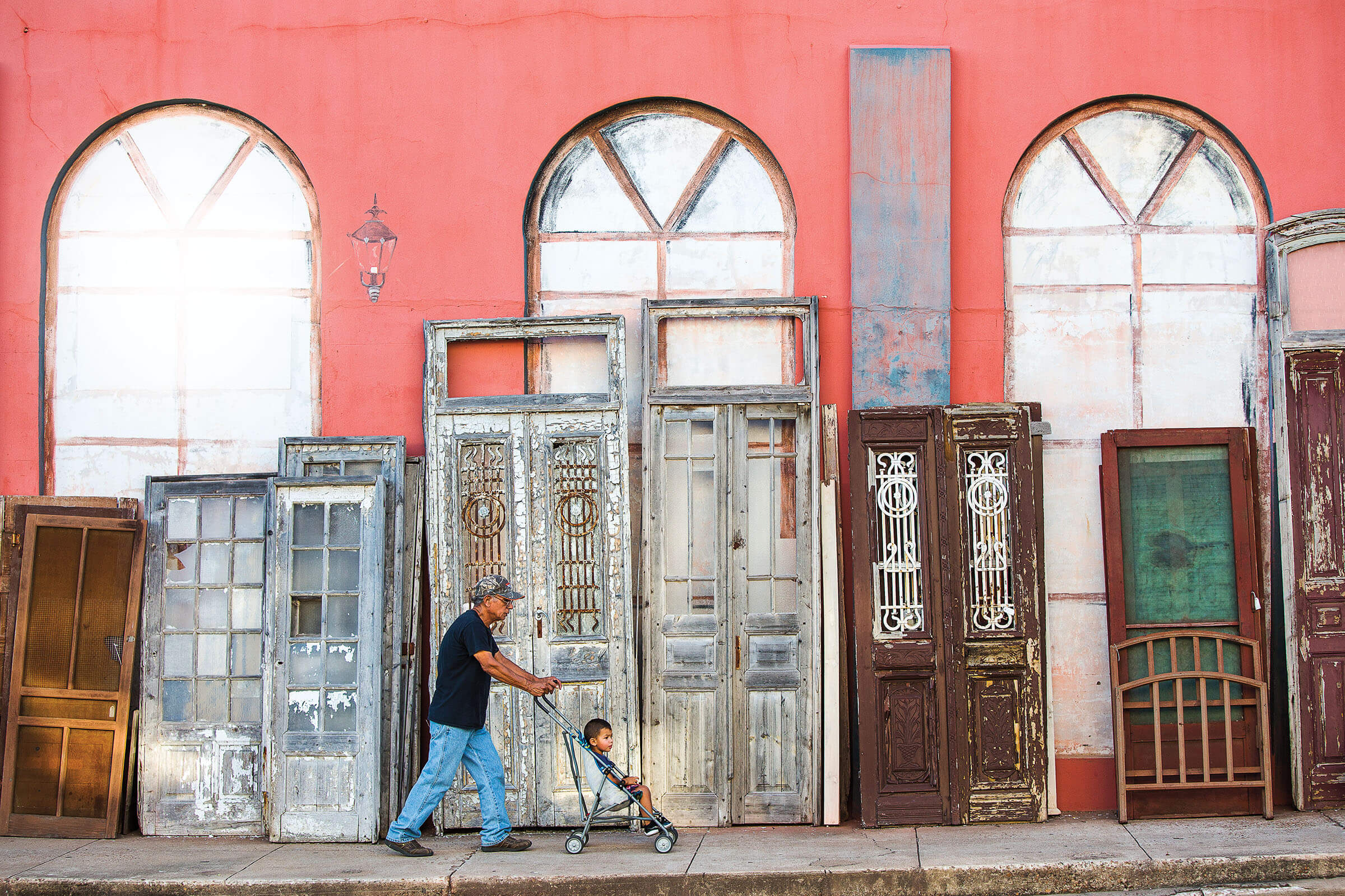 A man pushes a stroller in front of a pink wall with several antique doors in front in Brenham Texas