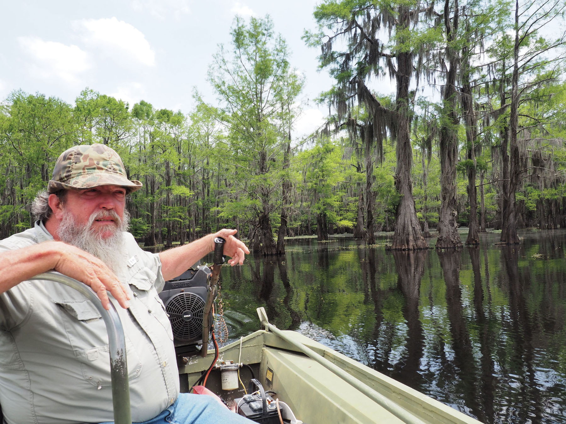 In a camoflauge hat and big beared, John Winn steers from the back of his boat tour on Caddo Lake Texas