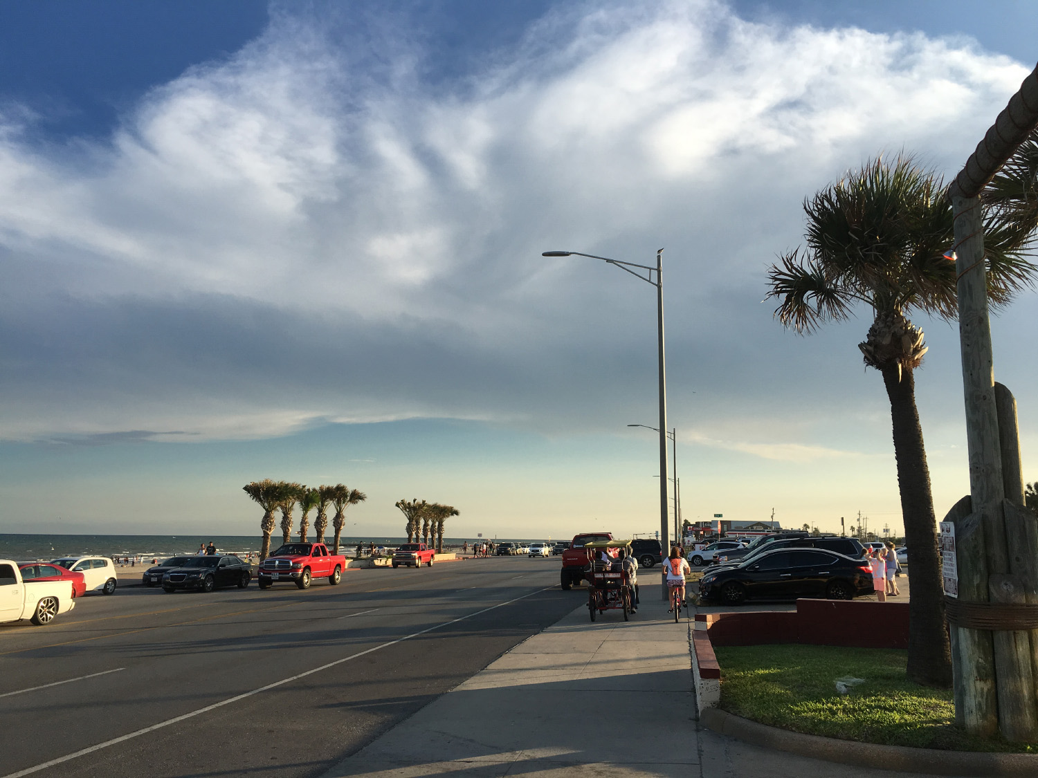 Clouds form above palm trees, parked cars and a bike rider along Seawall Boulevard on Galveston Island.