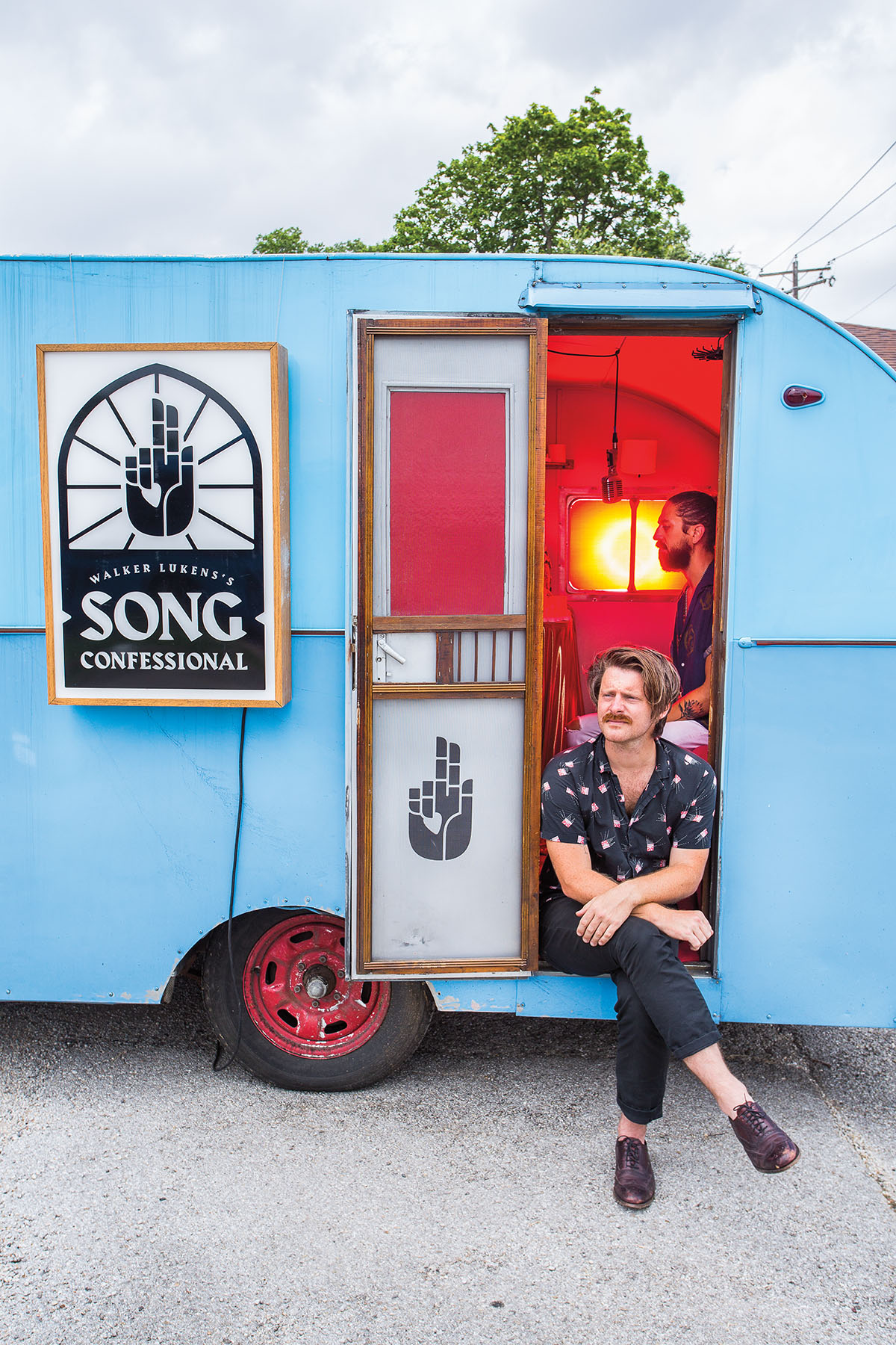 A man sits cross-legged in the doorway of a blue trailer with a sign reading "Walker Lukin's Song Confessional"
