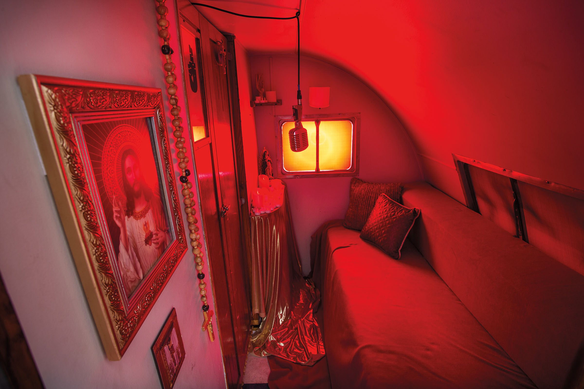 A microphone hanging from the ceiling is the focal point of the interior of the Walker Lukens Song Confessional, which is decorated with pieces of Catholic art as well as a rosary and glows with red light from the covered windows.