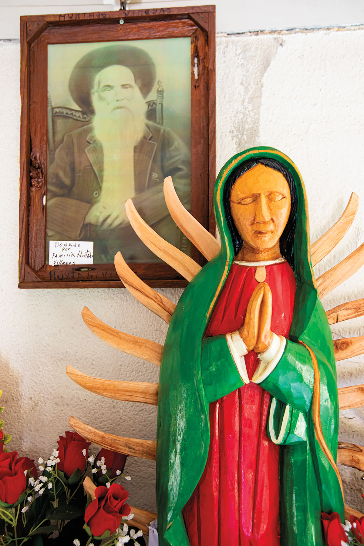 A wood-carved statue of Our Lady of Guadalupe dressed in a green robe and white dress, with a picture of Jaramillo in the background