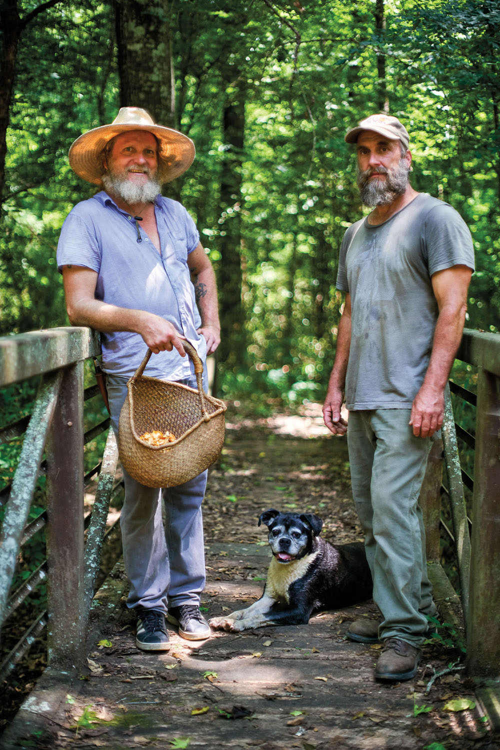 Two men in hats stand on a forested bridge with their grey and white dog