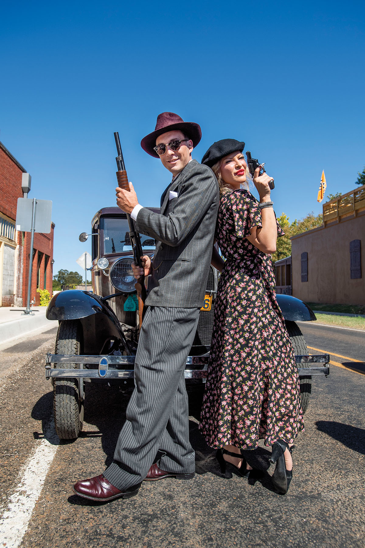 Two actors stand back to back in time-period clothes in front of an old car