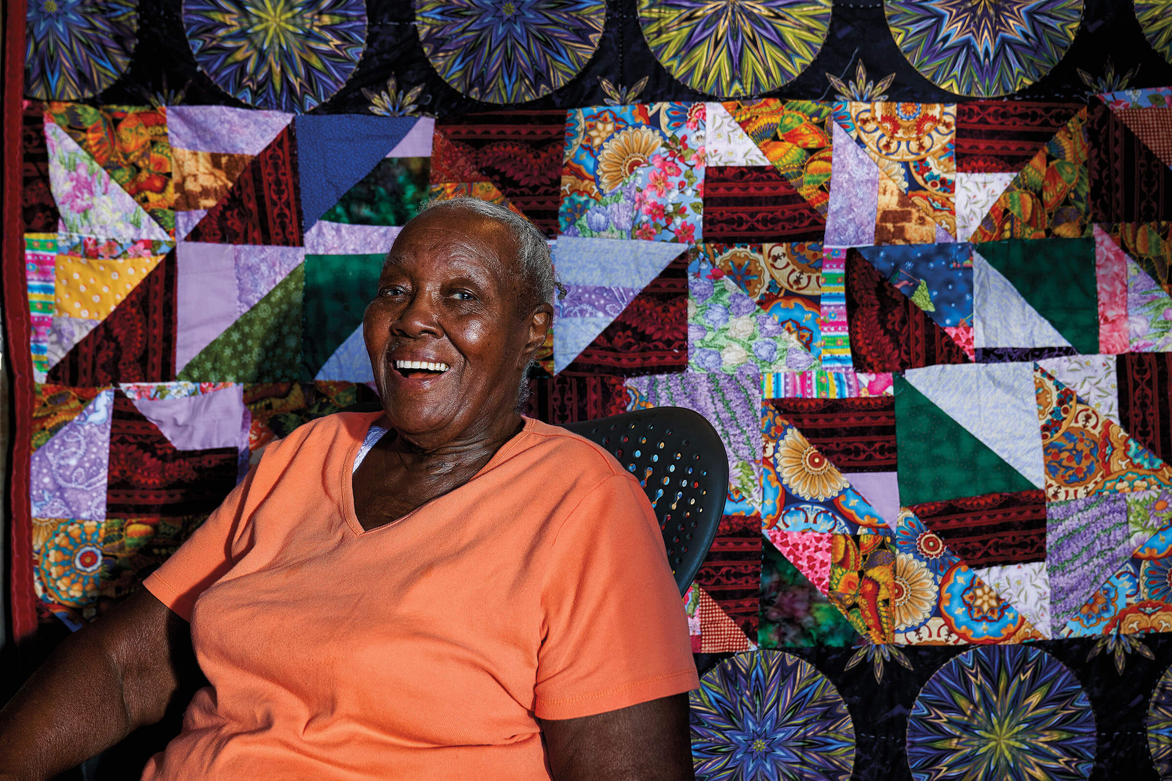 A picture of Laverne Brackens in an orange shirt smiling in front of a large quilt