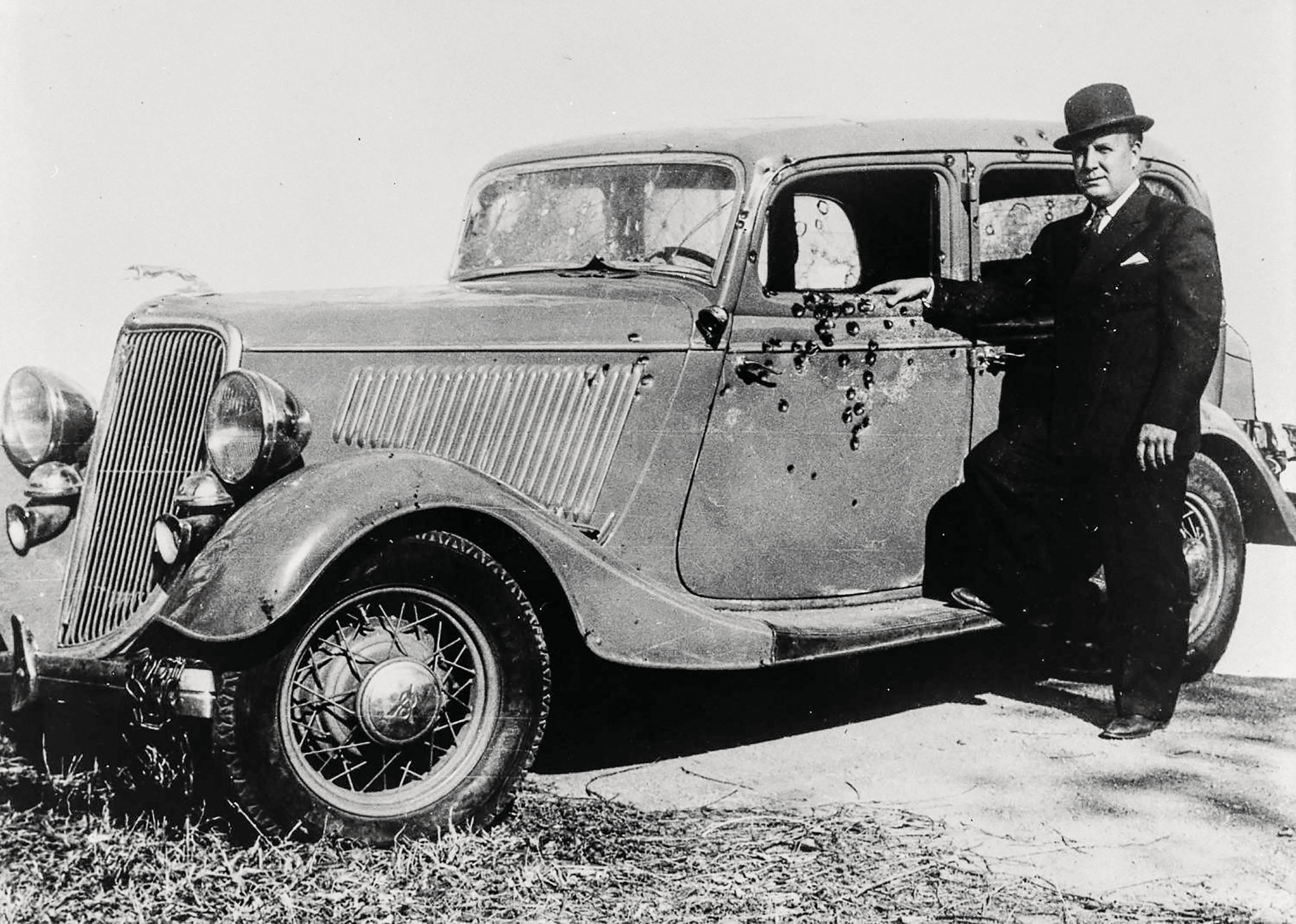 A man in a suit and hat stands next to Bonnie and Clyde's V-8 Ford covered in bullet holes