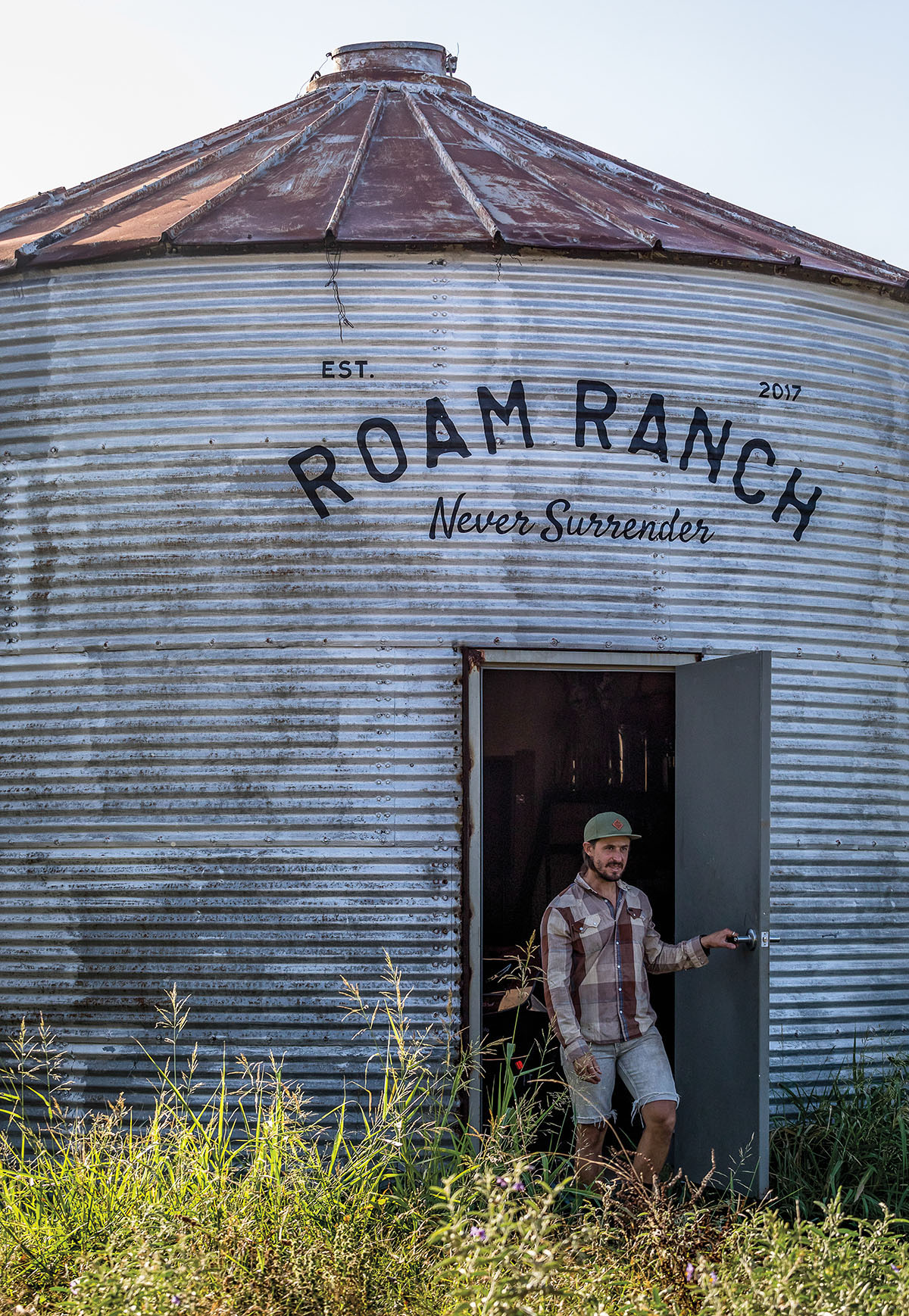 A man stands in the doorway of a rusted, silver silo reading "ROAM Ranch"