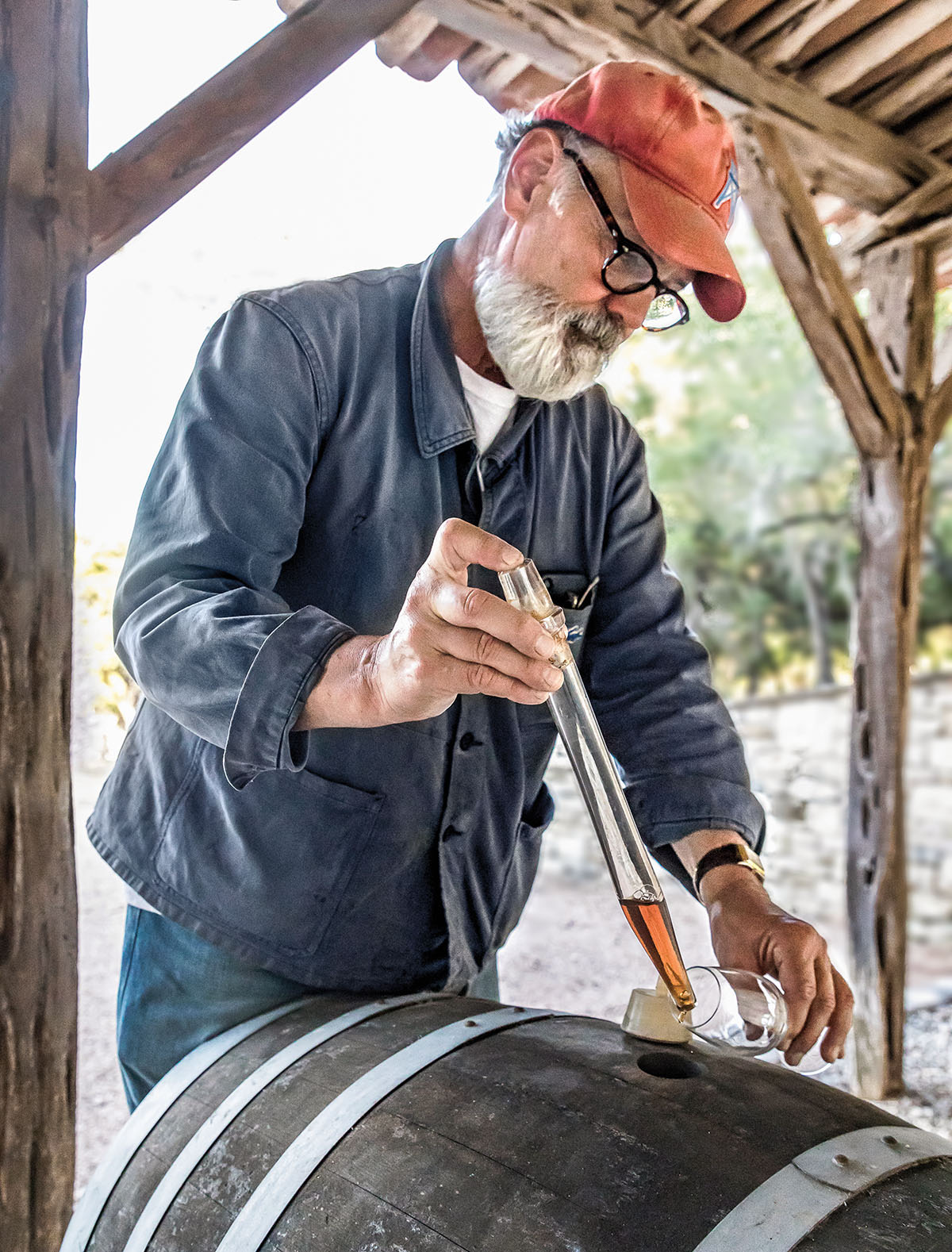 A man in an orange ball cap dips a testing device into a barrel of wine