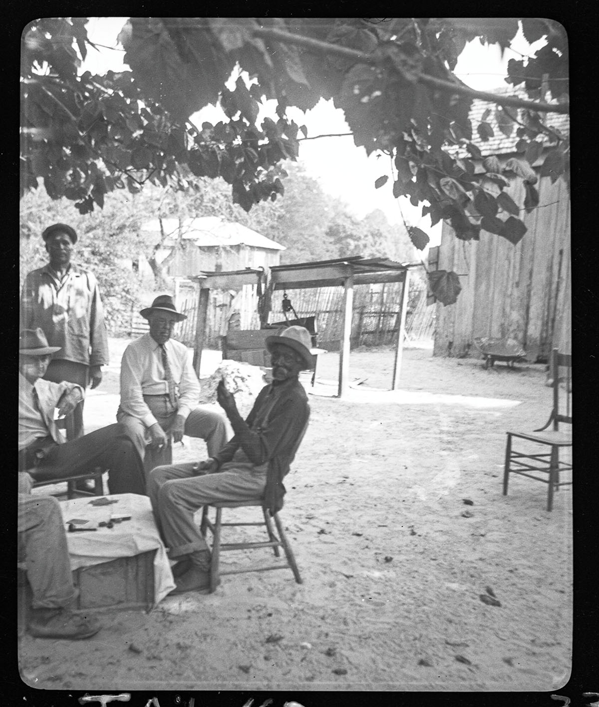 In this black and white photo, a group of men sit in wooden chairs on dirt ground