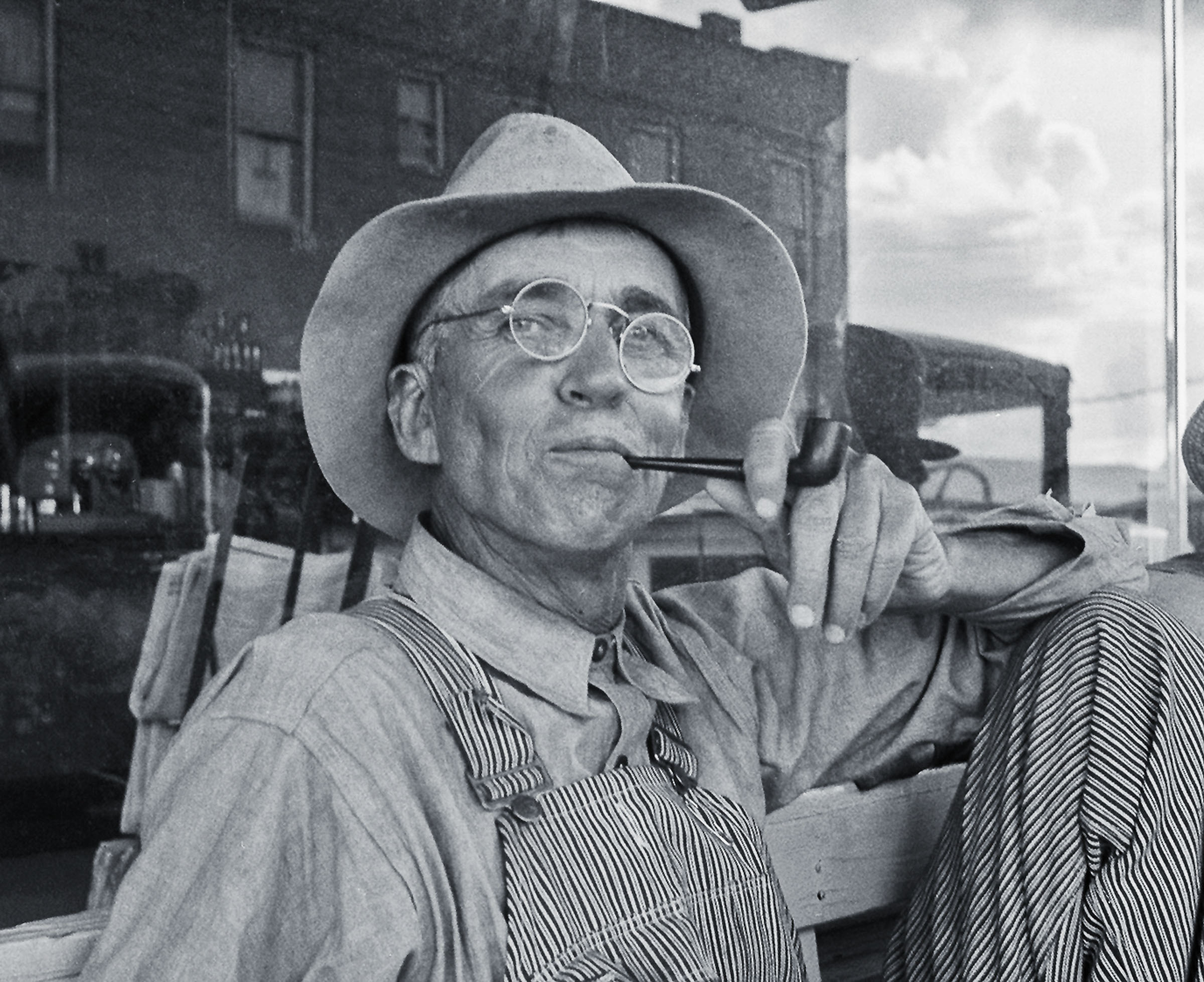 A man with a crooked wide-brimmed hat and crooked glasses smokes a piipe as a dust storm blows behind him