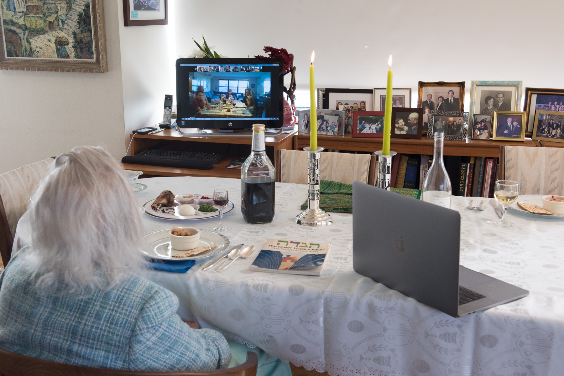 A woman with white hair sits alone at a table set for a Passover Seder meal. She looks at a screen with mutliple people on a Zoom call