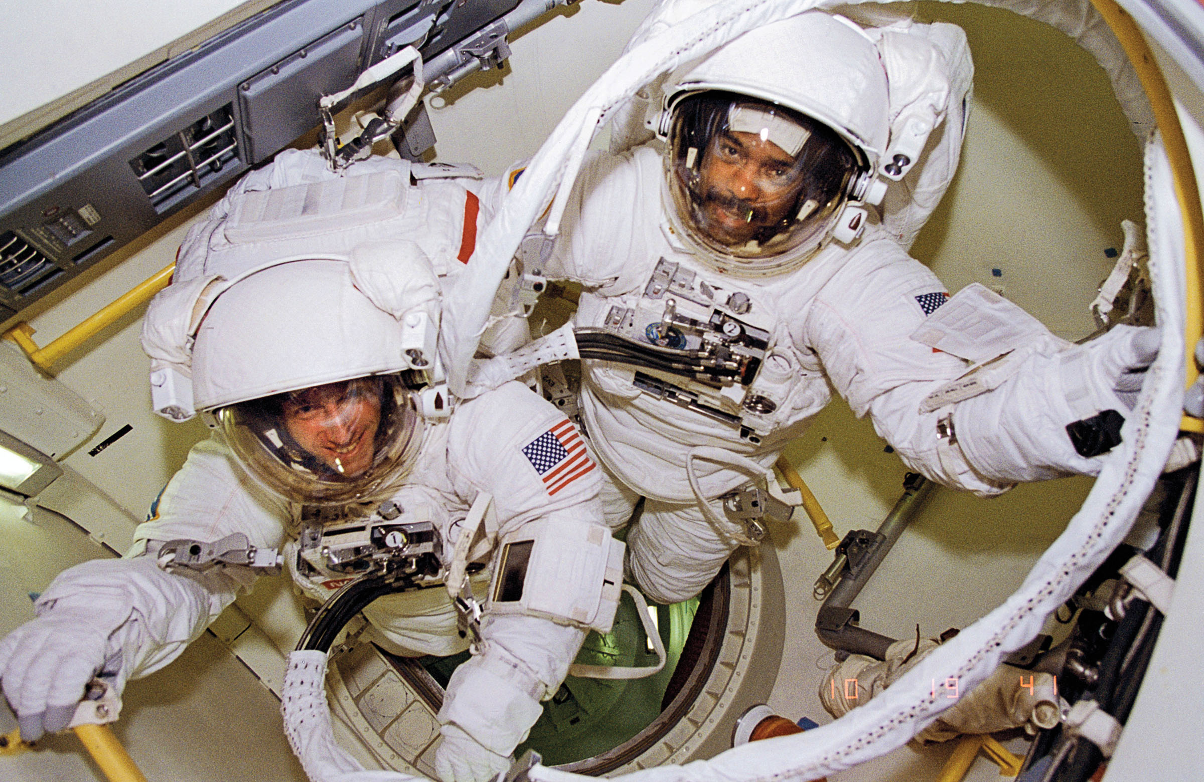 Astronauts in space suits inside of a capsule look up and smile at the camera