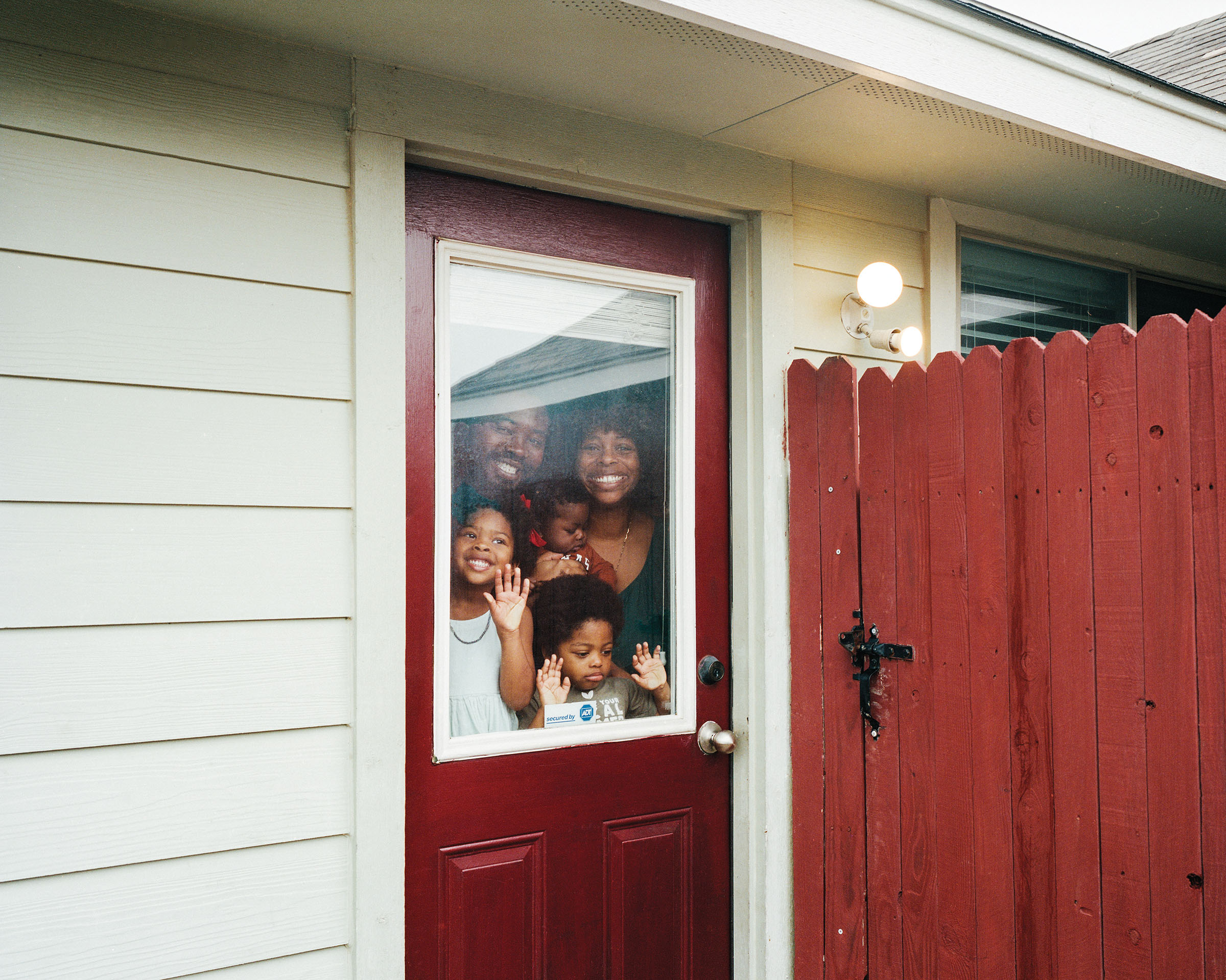 A family looks through the window in a dark red door next to a red fence