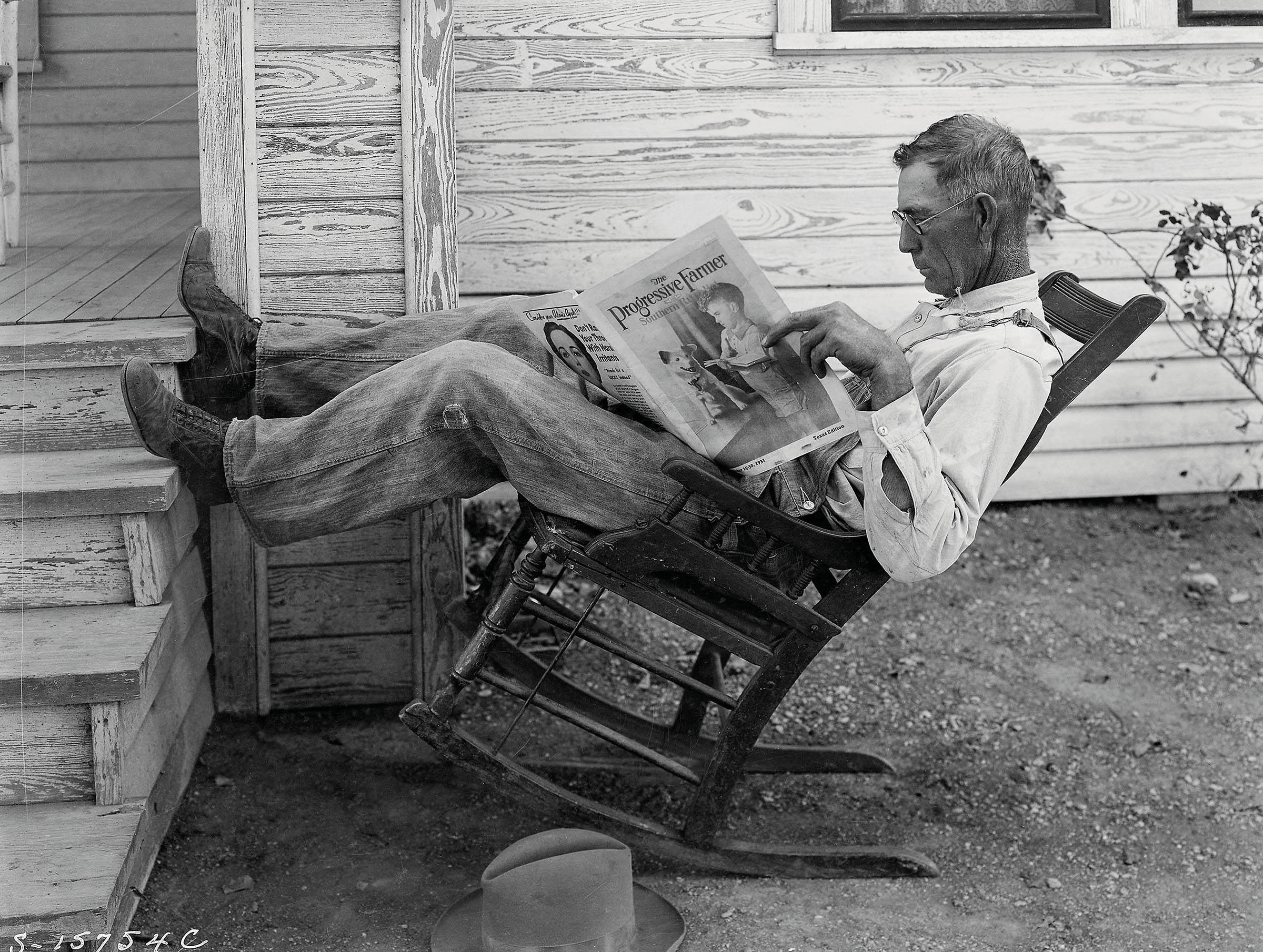 A man leans back in a rocking chair while reading a newspaper in this historic photo