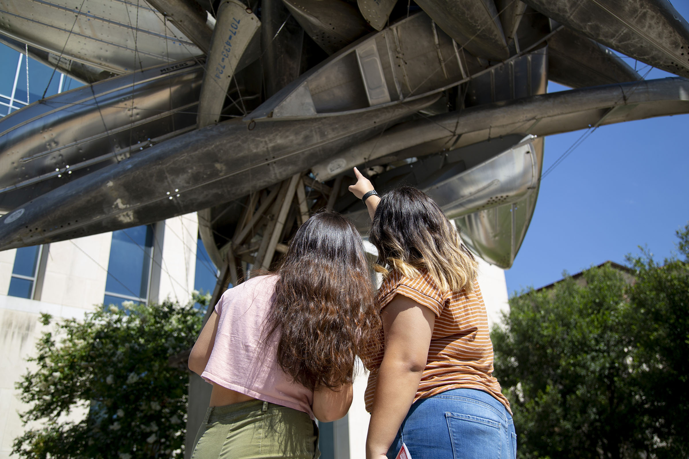 Two women point at a gray metal sculpture from below