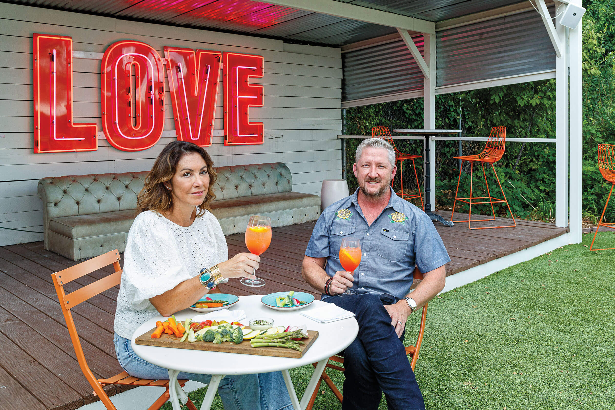 A couple sits at a folding table outside of a building with a large neon sign reading "LOVE"