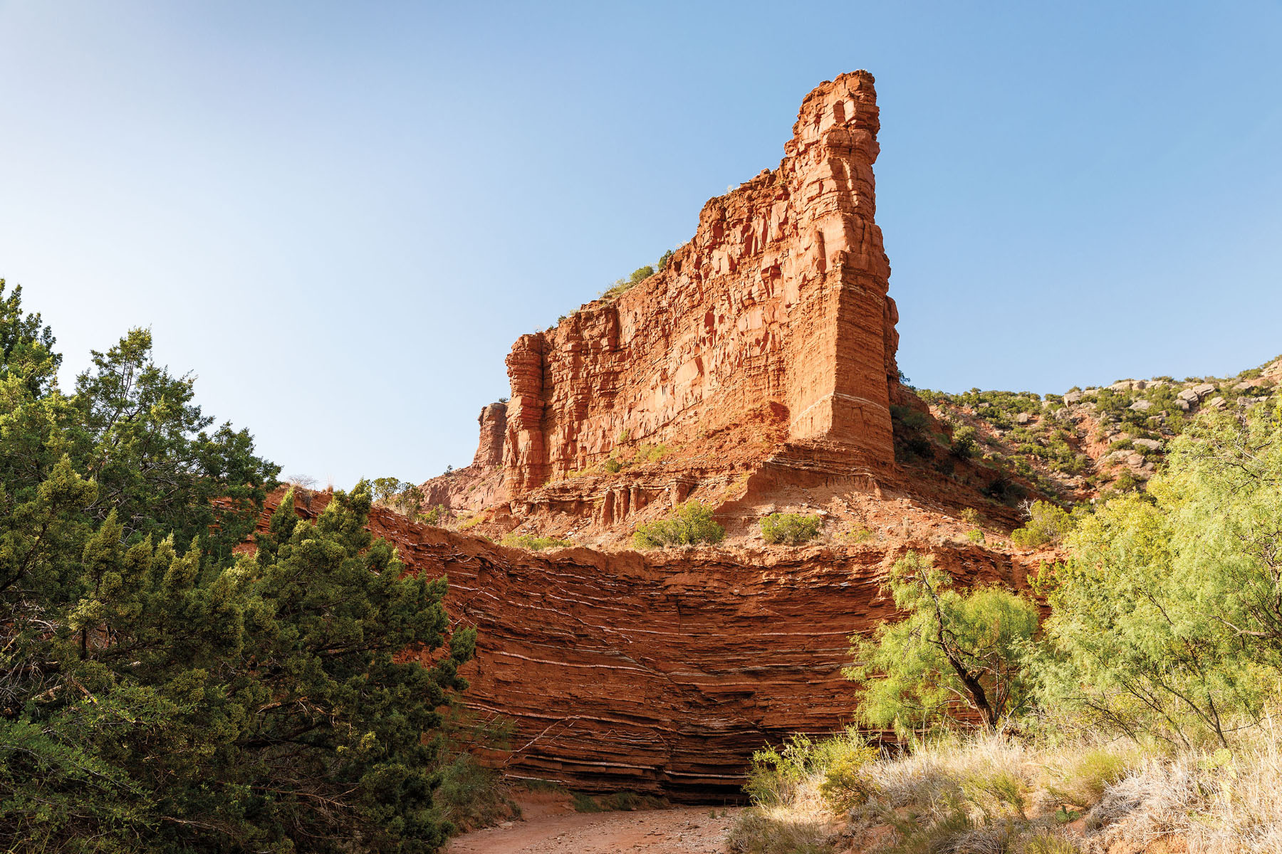 A red rock pillar surrounded by scrubby green trees and sedimentary rock