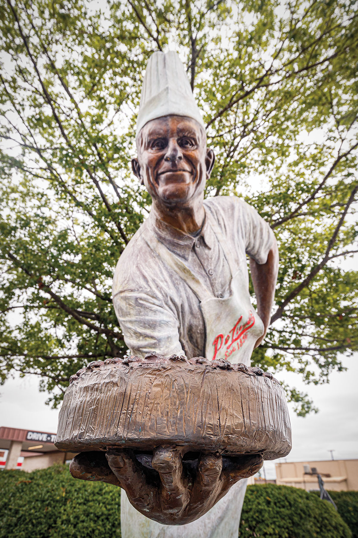 A statue of a man in a white chef's hat and apron holding a bronze fruitcake