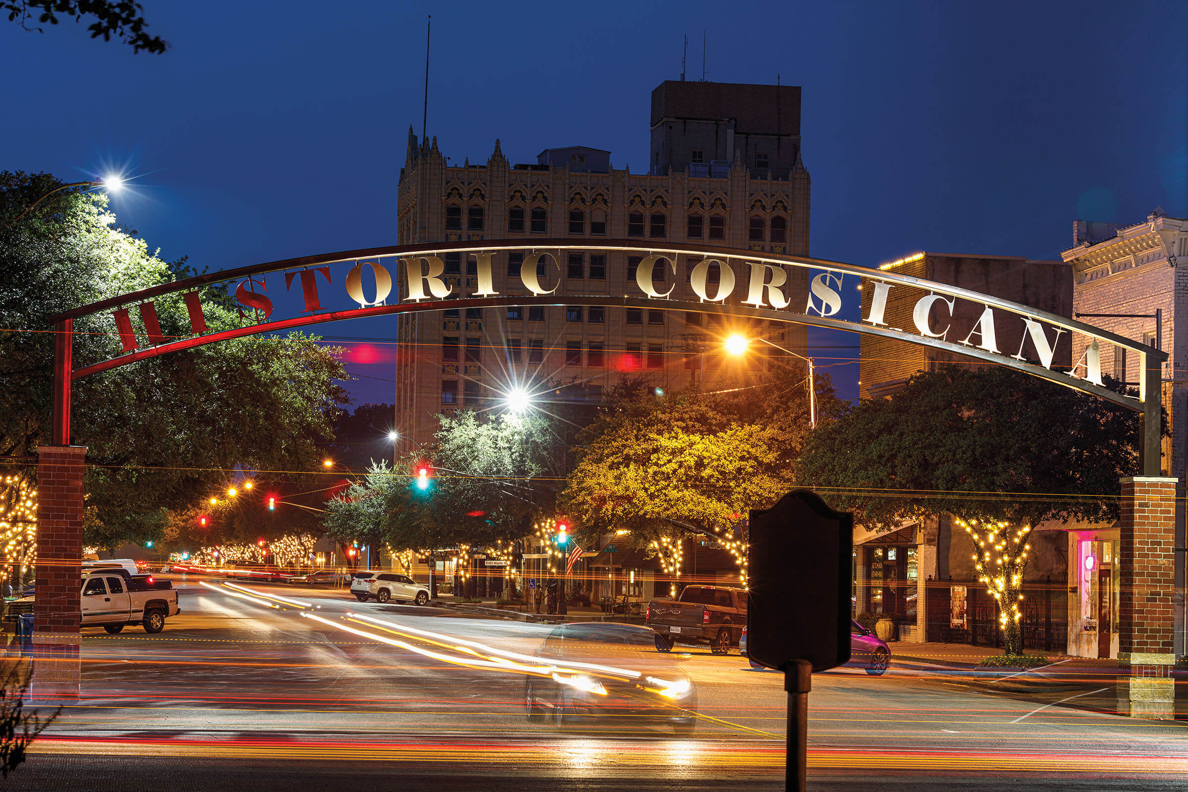 A nighttime view of an iron arch reading "Historic Corsicana" with the glow of car headlights and taillights