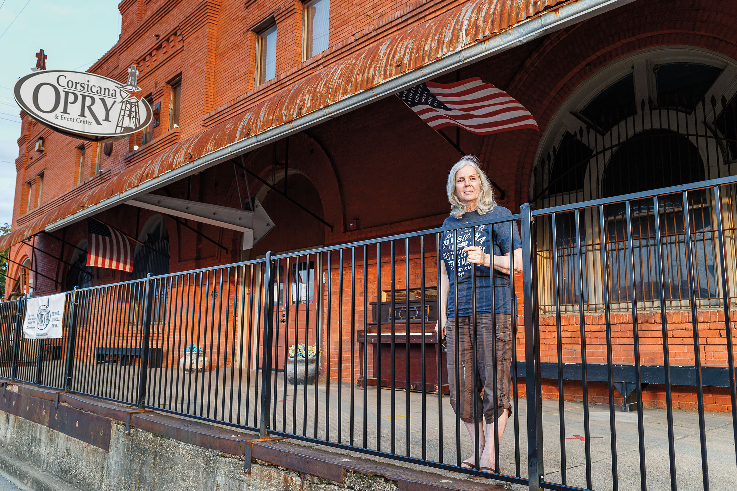 A woman stands on a concrete slab behind an iron fence under a sign reading "Corsicana Opry"