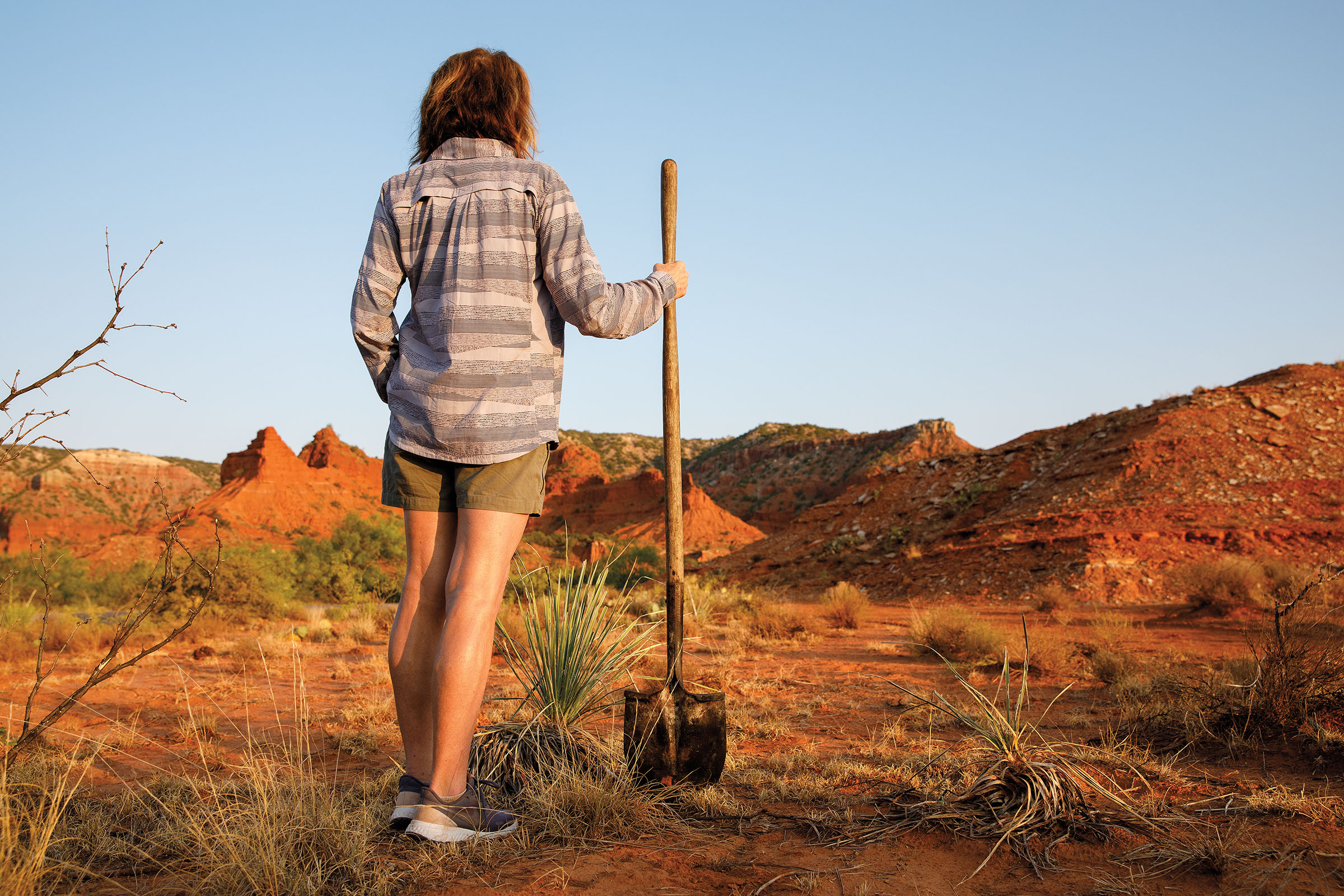 A woman stands with a shovel next to several plants in the desert