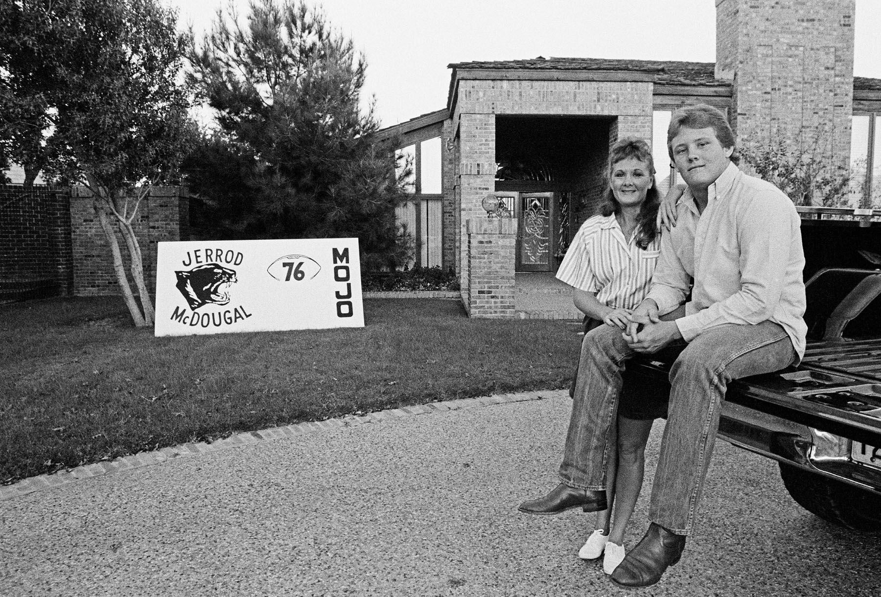 A young man and his mother sit on the tailgate of a truck next to a sign reading "Jerrod McDougal, 76, MOJO"