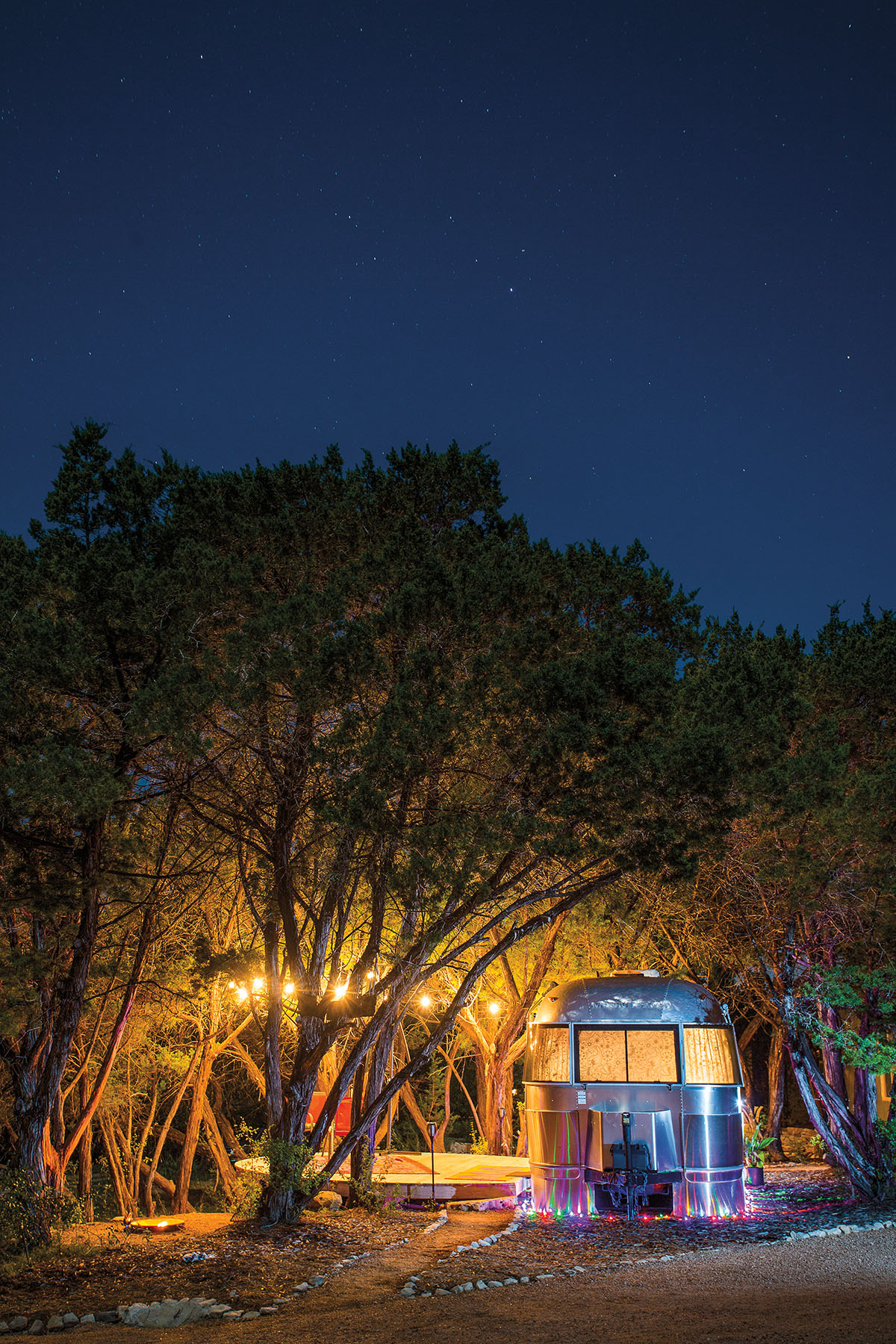 An Airstream trailer with string lights underneath a canopy of trees at night