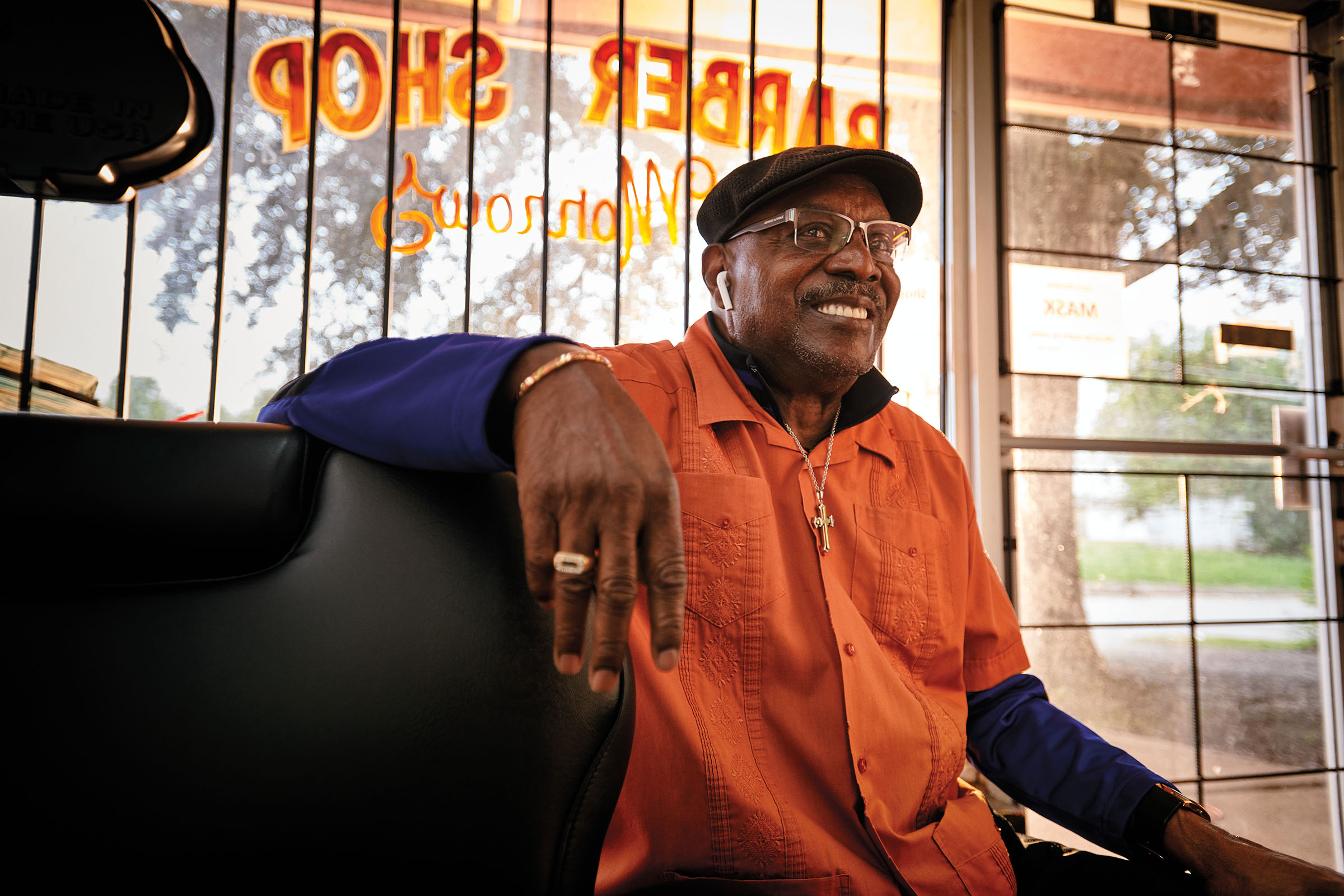 A man in an orange shirt smiles from a chair in front of the window of his barber shop