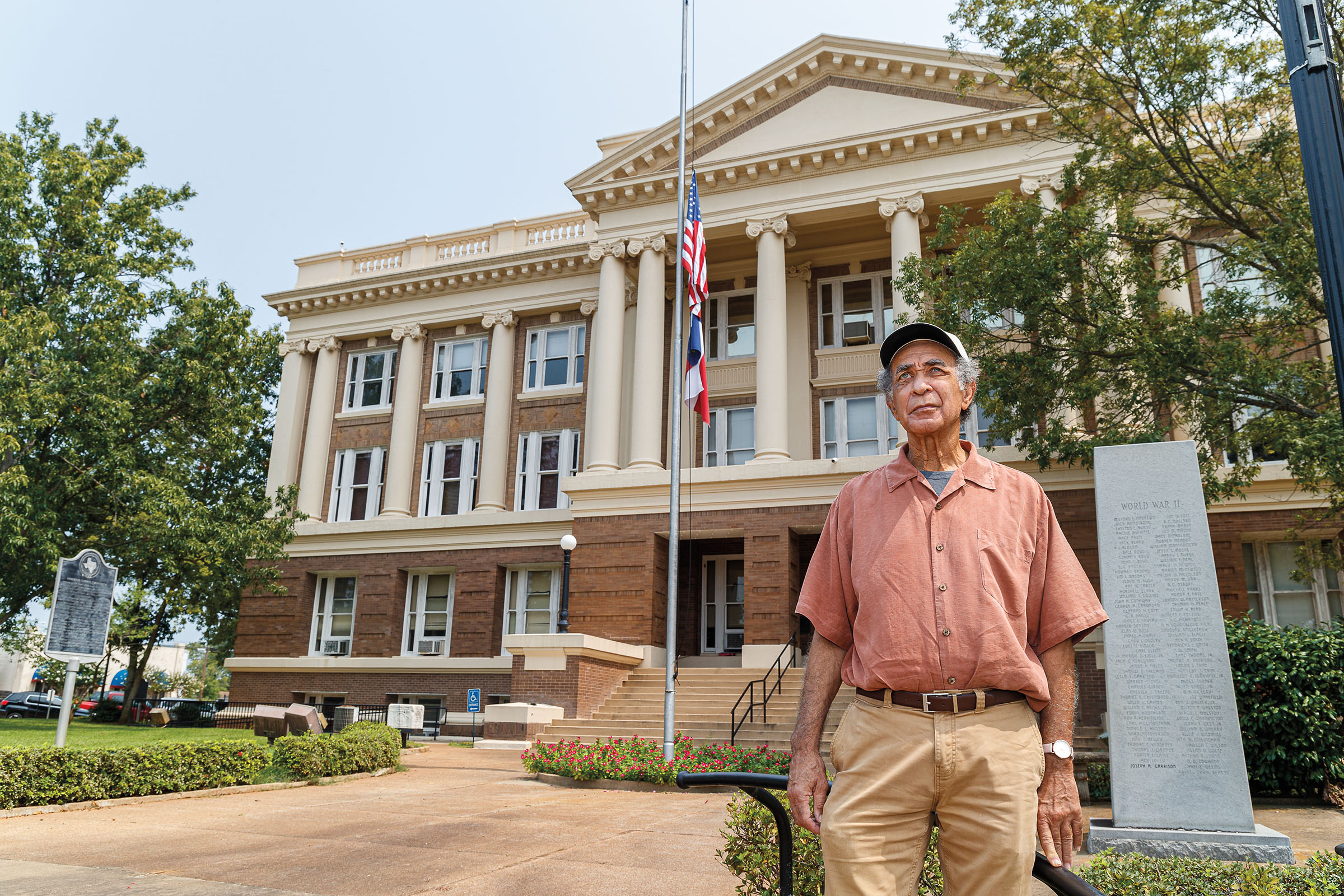 A man in a button-down shirt stands in front of a courthouse with a Texas and United States flag