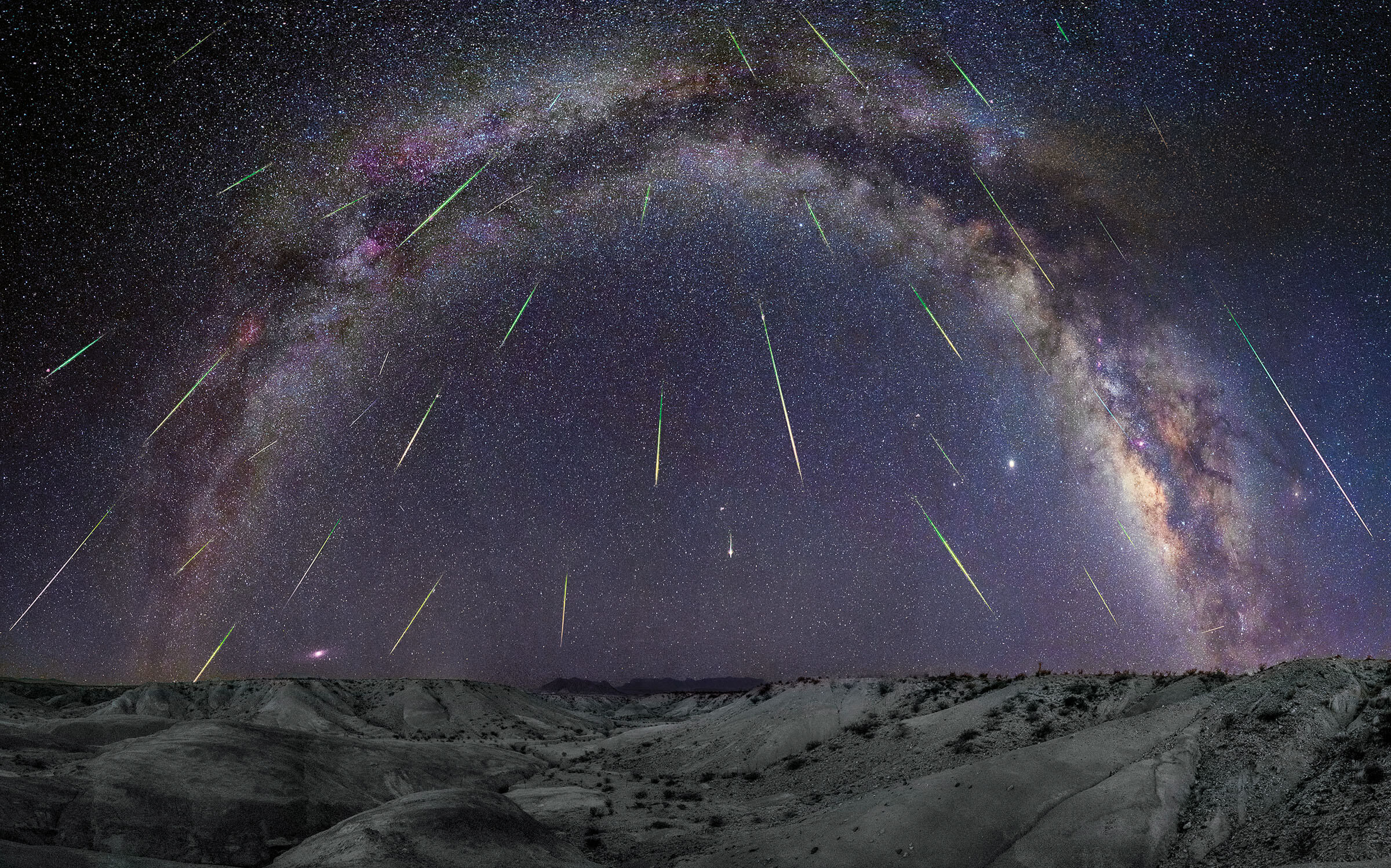 An arc of the Milky Way galaxy with meterors streaming down above gray sand desert