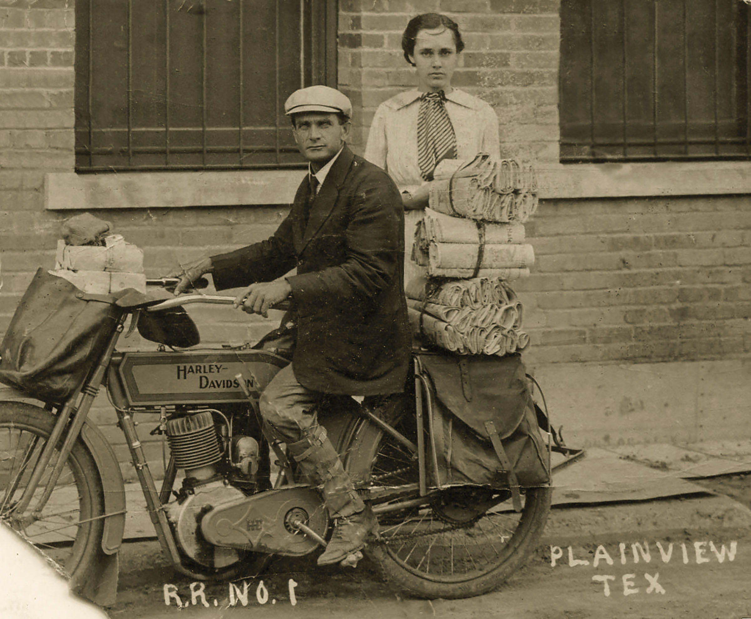 A sepia-tone photograph of a man on a motorcycle and a woman behind him. The motorcycle is loaded down with letters and packges.