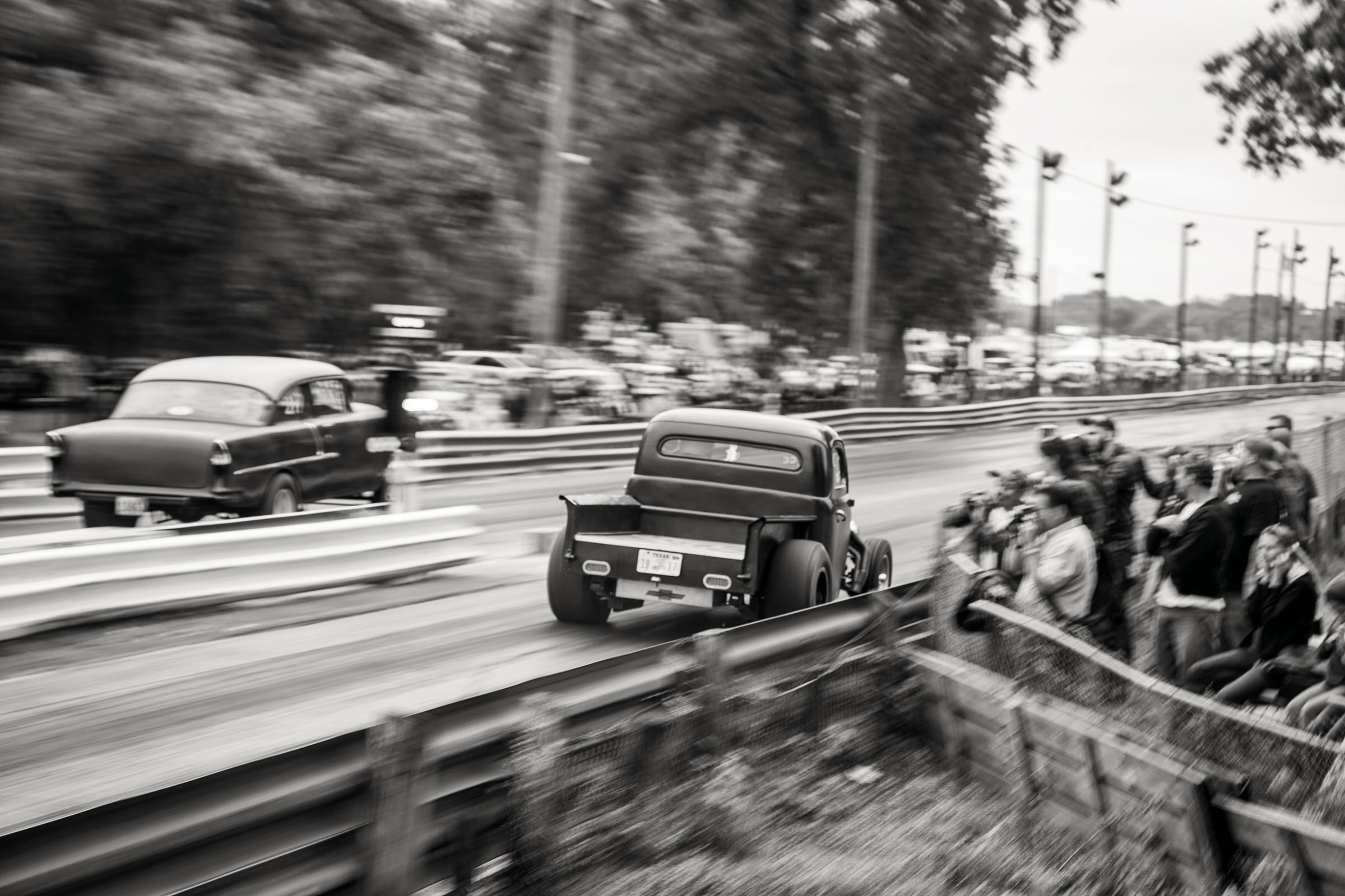 Two cars race down a road with guardrails as a crowd looks on 