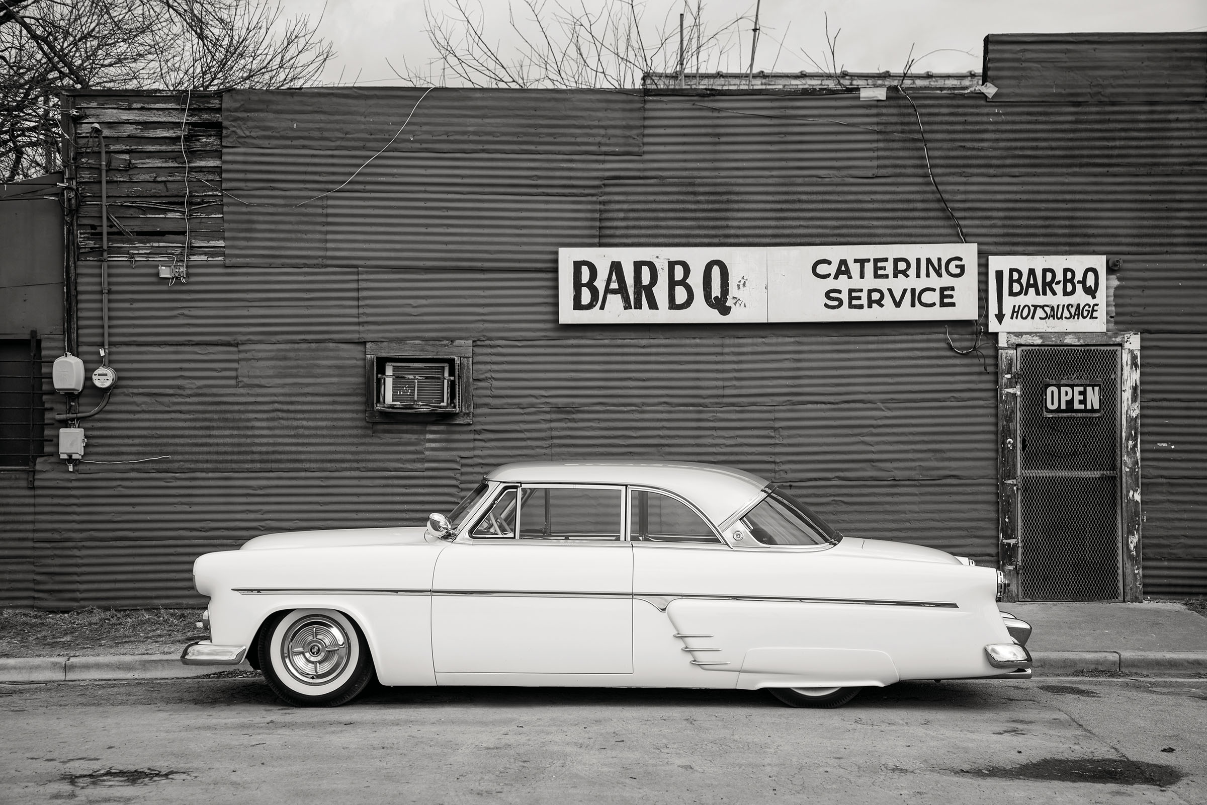 A long, white car in front of a brick building with a sign reading "Bar BQ Catering Service"