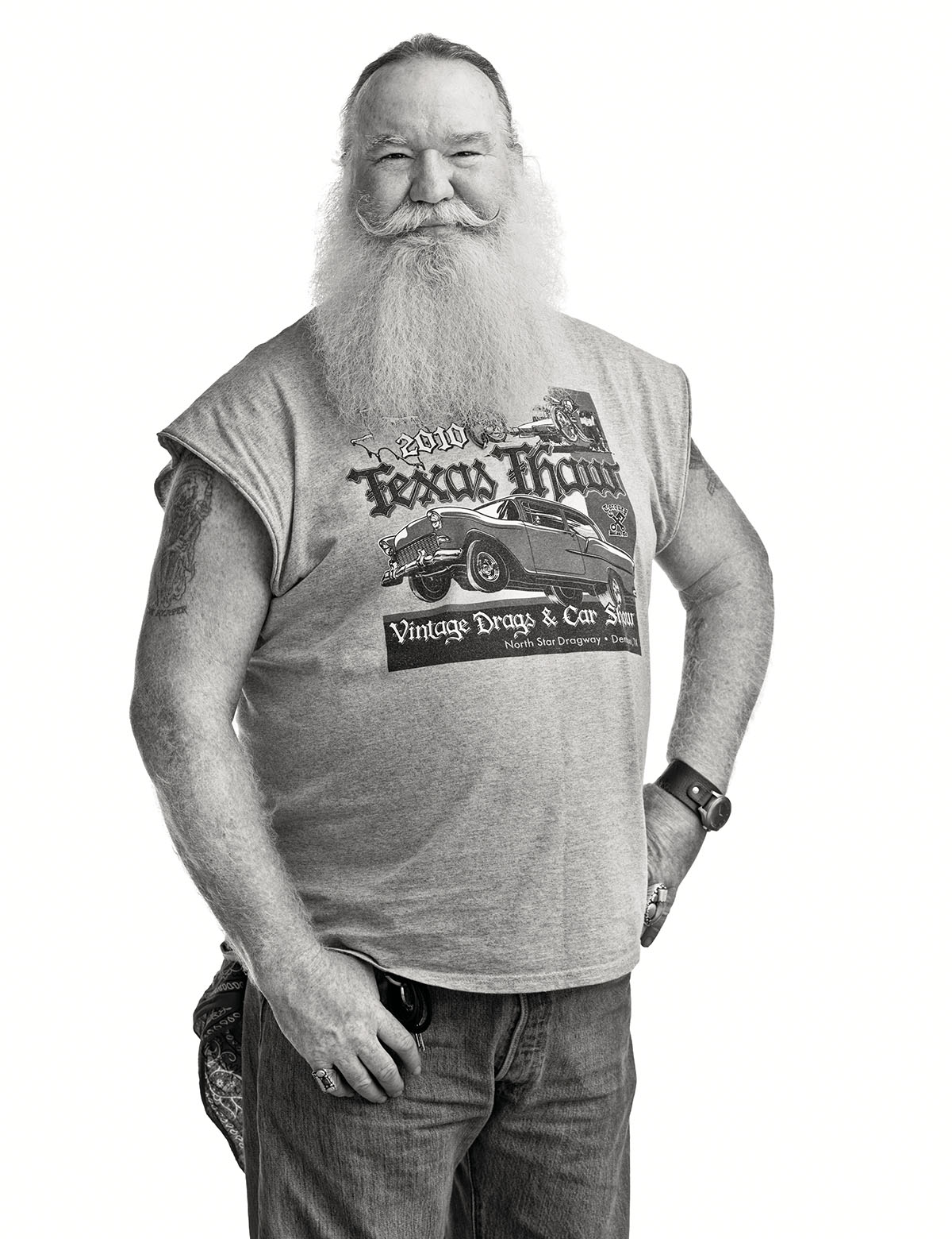 A man with a large white beard in a cut-off t-shirt stands with his hands in his pockets