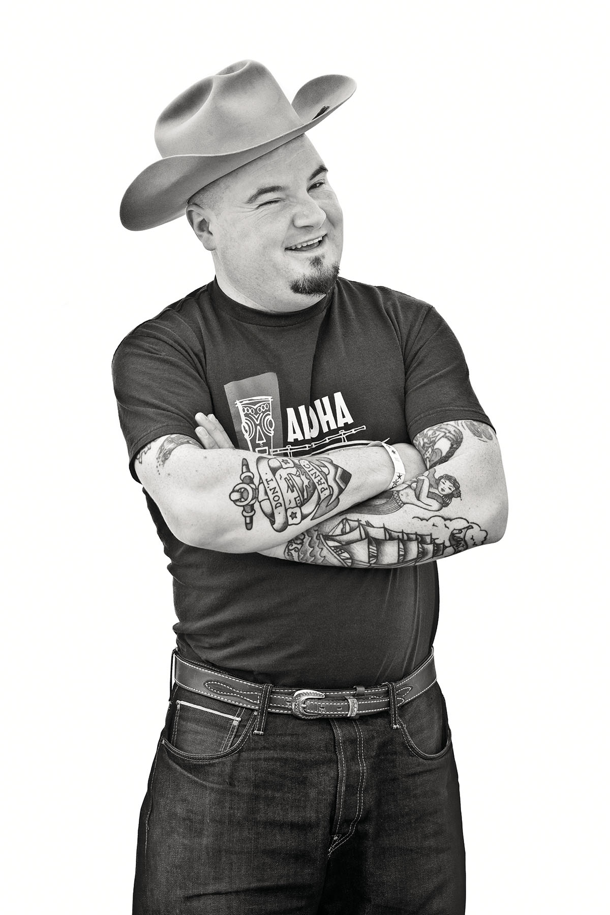 A man in a cowboy hat and black t-shirt crosses tattooed arms