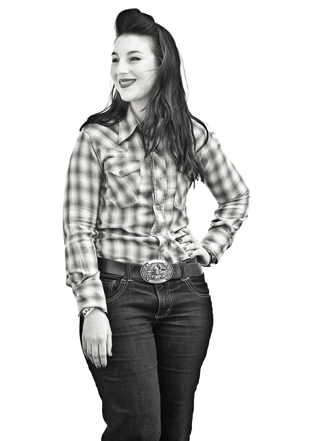 A woman in a plaid shirt and jeans with a large belt buckle poses with her hand on her hip