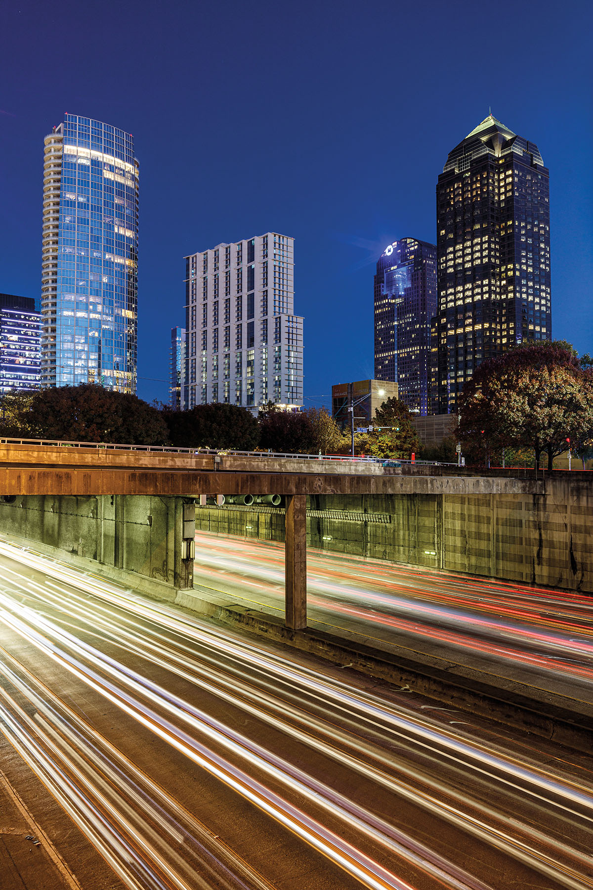 Lights stream from passing vehicles on a freeway below buidlings lit up in downtown Dallas