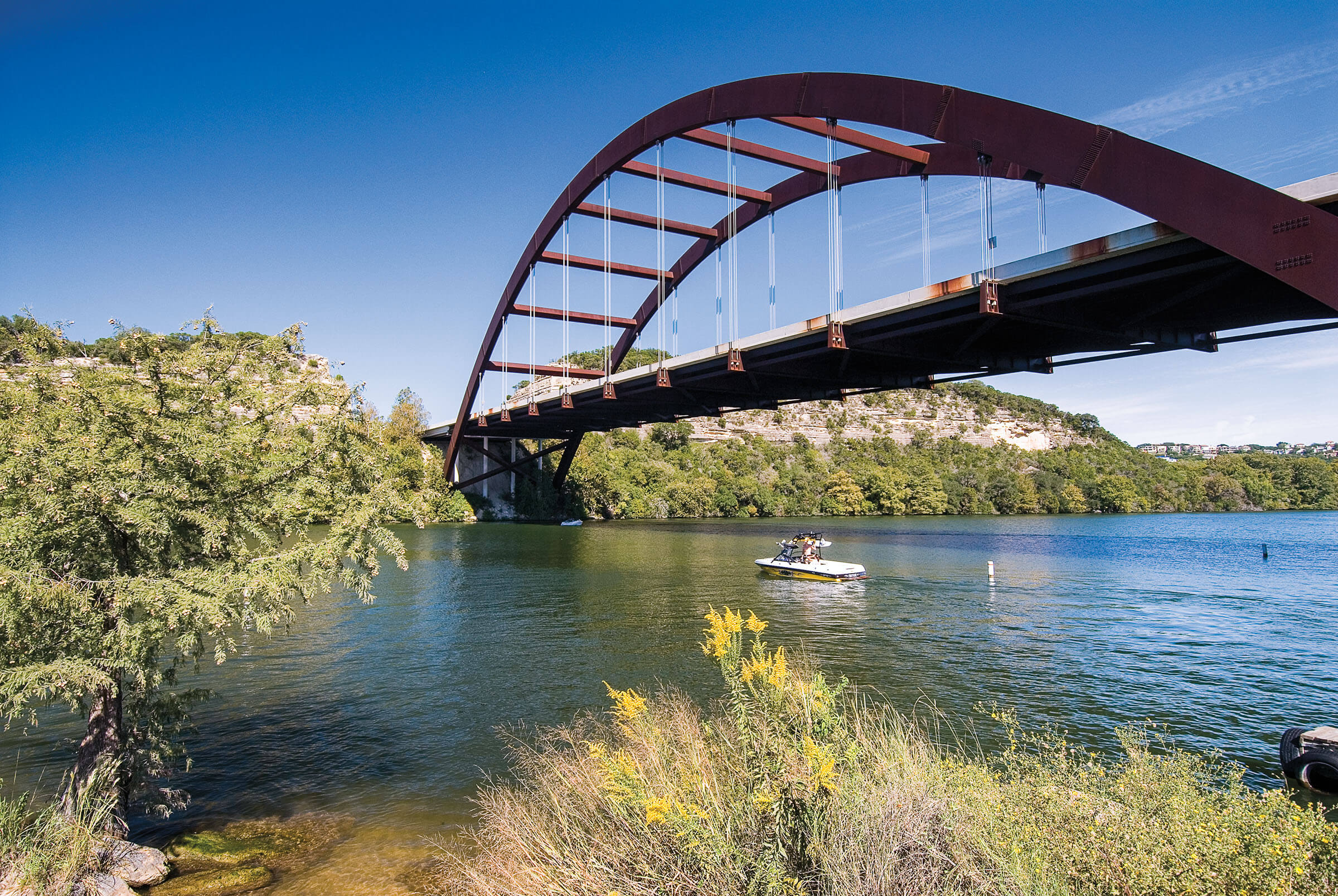 A photo from the shoreline of the Pennybacker Bridge in Austin, with blue sky, blue water and a yellow motorboat