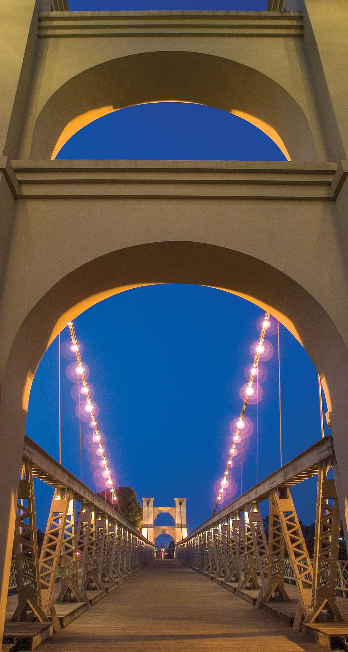 A view upward from the Waco Suspension Bridge showing a dark blue night sky and purple lights illuminating the bridge