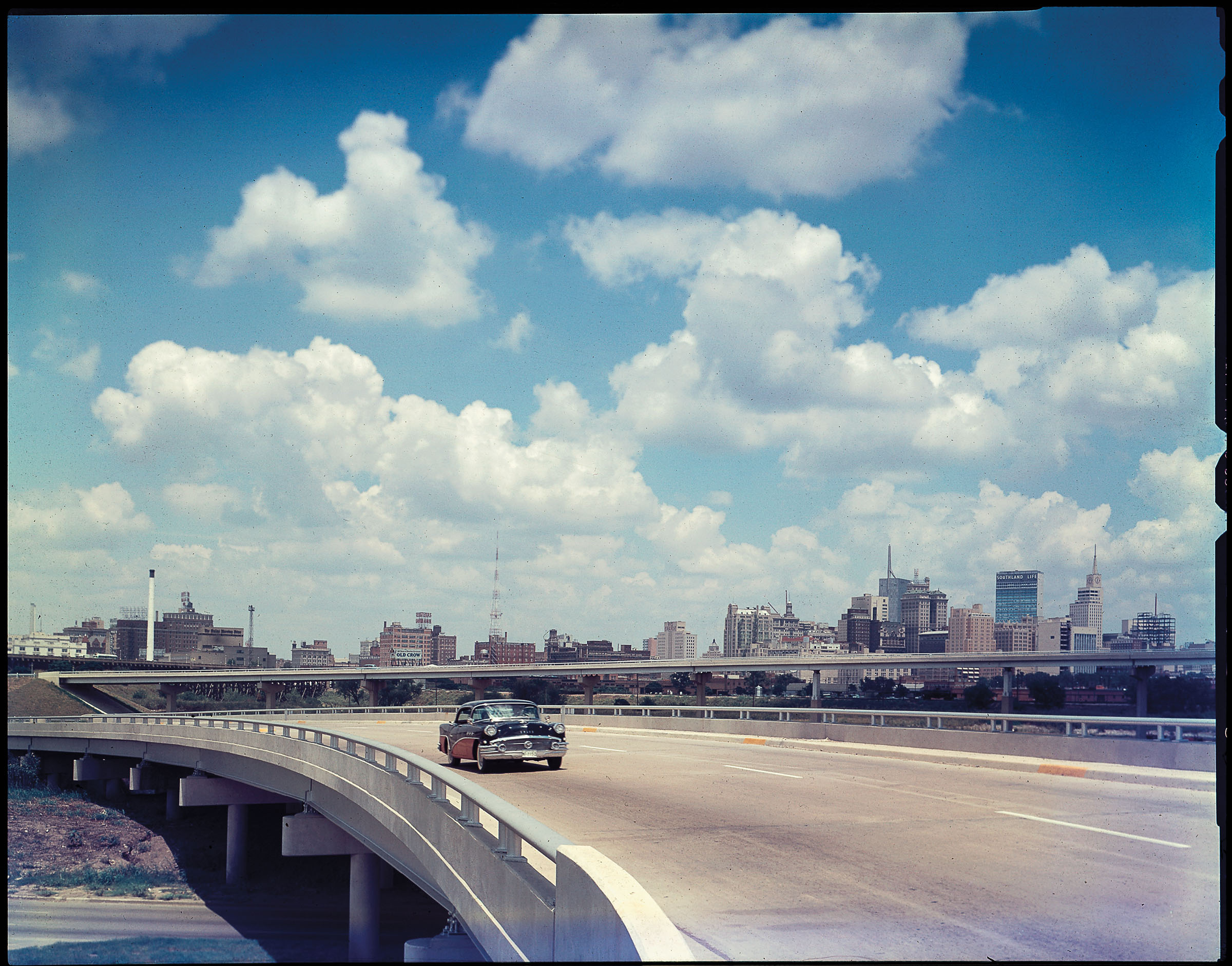 A vintage photo of a car driving on a bridge with the Dallas skyline in the background