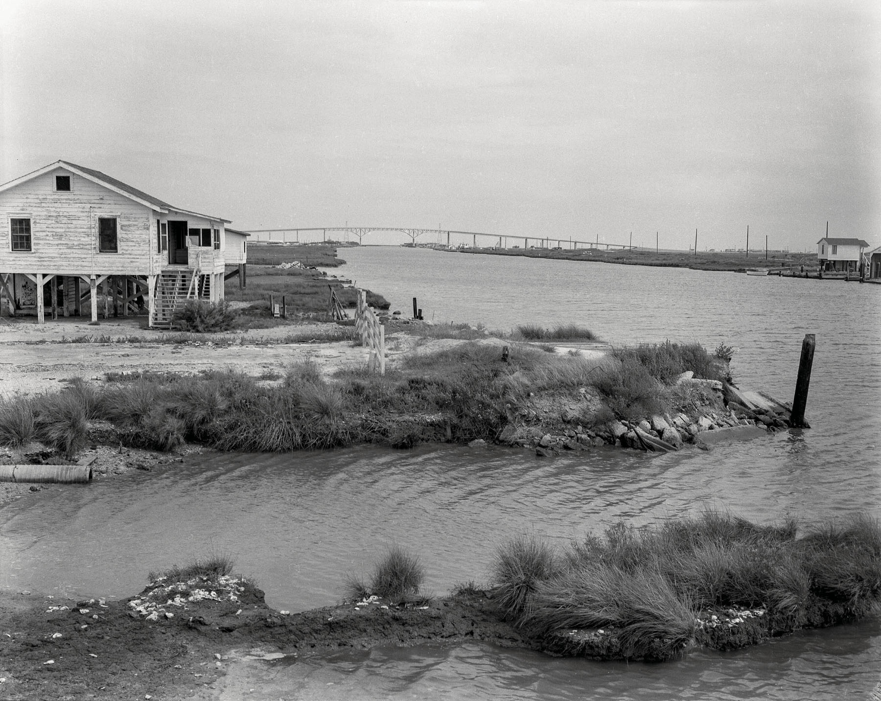 A white house with stairs on the beach, with a view of water extending into a small bay and a bridge in the distance