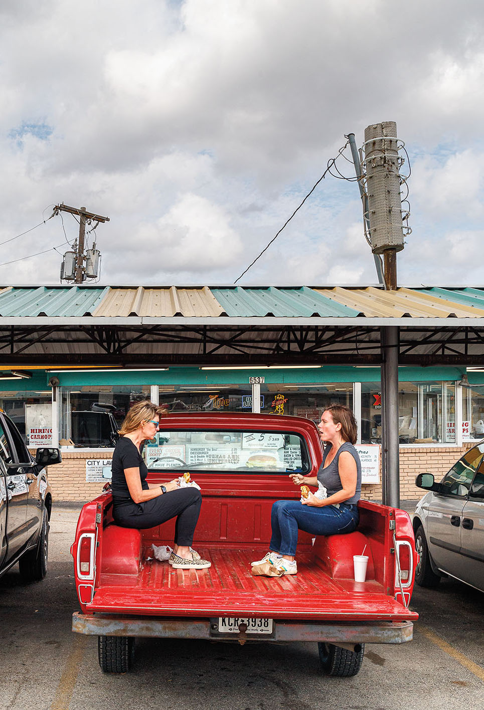 Two woman sit in the back of a red puckup truck outside of a store