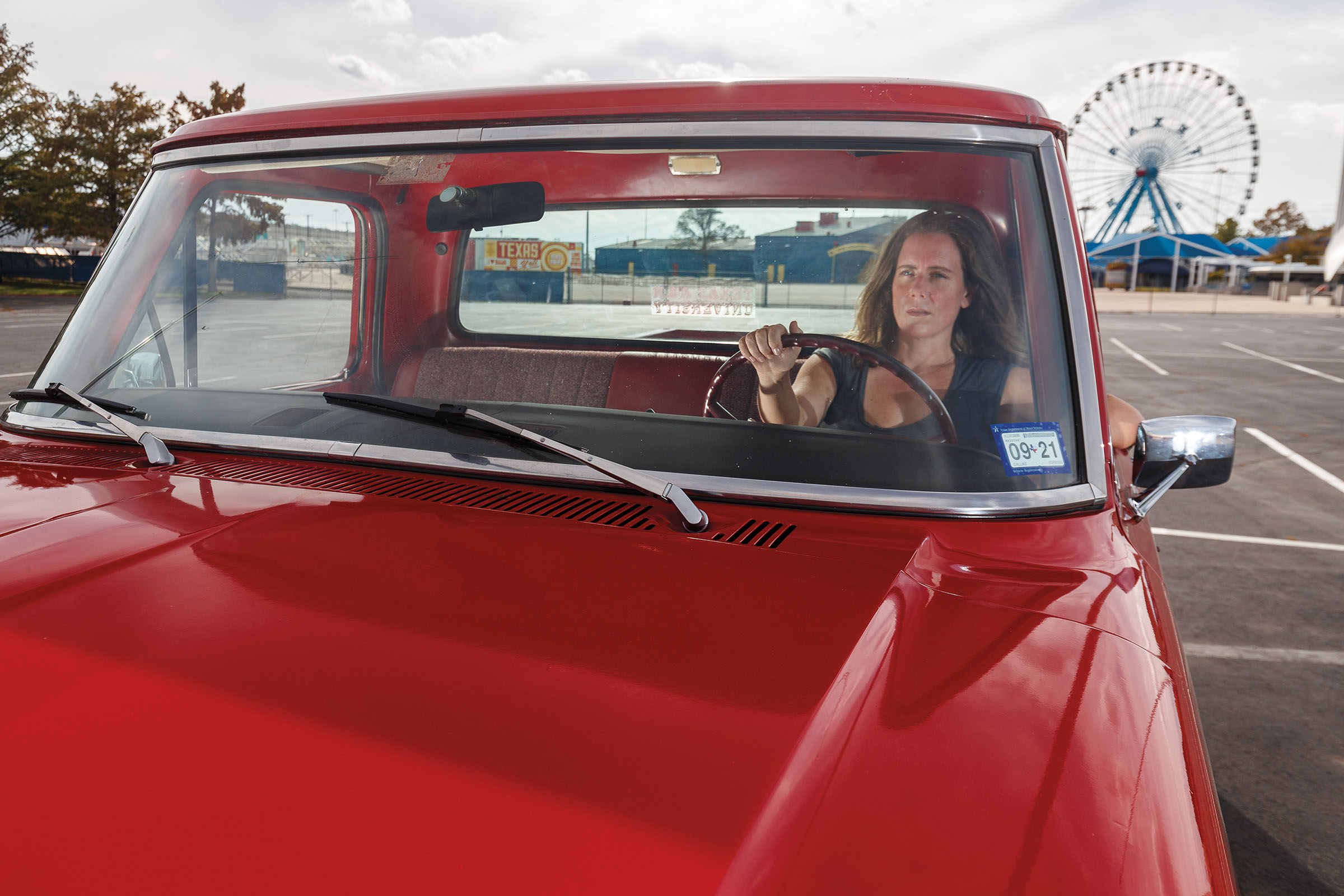 A woman drives a bright red Ford truck with the ferris wheel of Dallas' Fair Park in the background