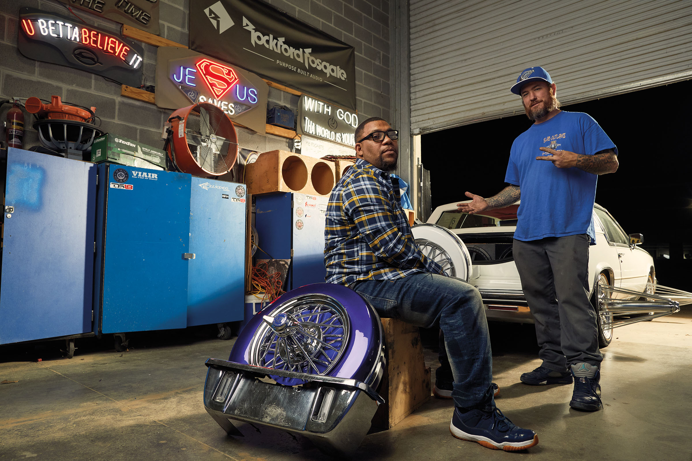 Two men stand in a garage surrounded by neon lights, industrial cabentry and shiny car wheels